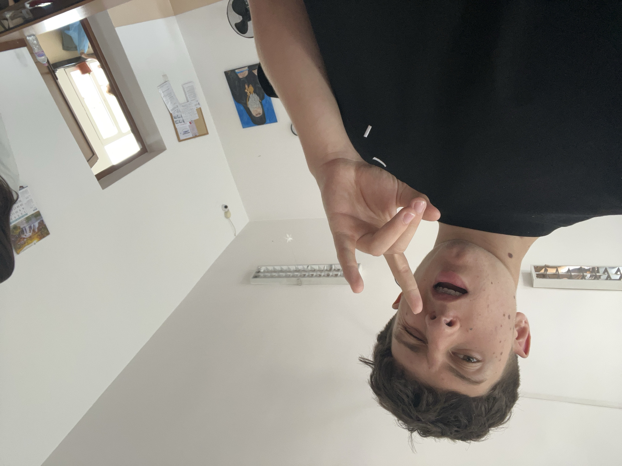 The image, taken in Zlatograd, Bulgaria, shows a young male in an indoor setting, captured from a low angle and presented upside down. The subject has short, dark brown hair and a fair complexion with numerous freckles across his face, particularly on his nose and cheeks. A mole is visible on his neck, just below his right ear. He is wearing a plain black t-shirt. His expression is playful, featuring a wink of his right eye (viewer's left) and a slightly open mouth. His left hand is raised, making a "V" or peace sign gesture with his index and middle fingers extended.
The background consists primarily of white or off-white walls. To the left of the person's head (when correctly oriented), a window or doorway opening reveals a brightly lit area beyond, suggesting it is daytime. A plastic water bottle rests on a ledge near this opening. Above the window, a dark-colored shelf or piece of furniture is partially visible. On the wall to the right, a black, three-bladed ceiling fan and a small cork bulletin board with several pieces of paper attached are visible. Below the fan, there's a framed artwork depicting a person with a blue background. A long, rectangular fluorescent light fixture is mounted on the ceiling, behind the subject's head. Part of a calendar or picture showing a landscape with a river or waterfall is visible on the far left wall. A small, white, wall-mounted device is also present below the bulletin board. No legible text is discernible in the image. The overall mood is informal and casual.