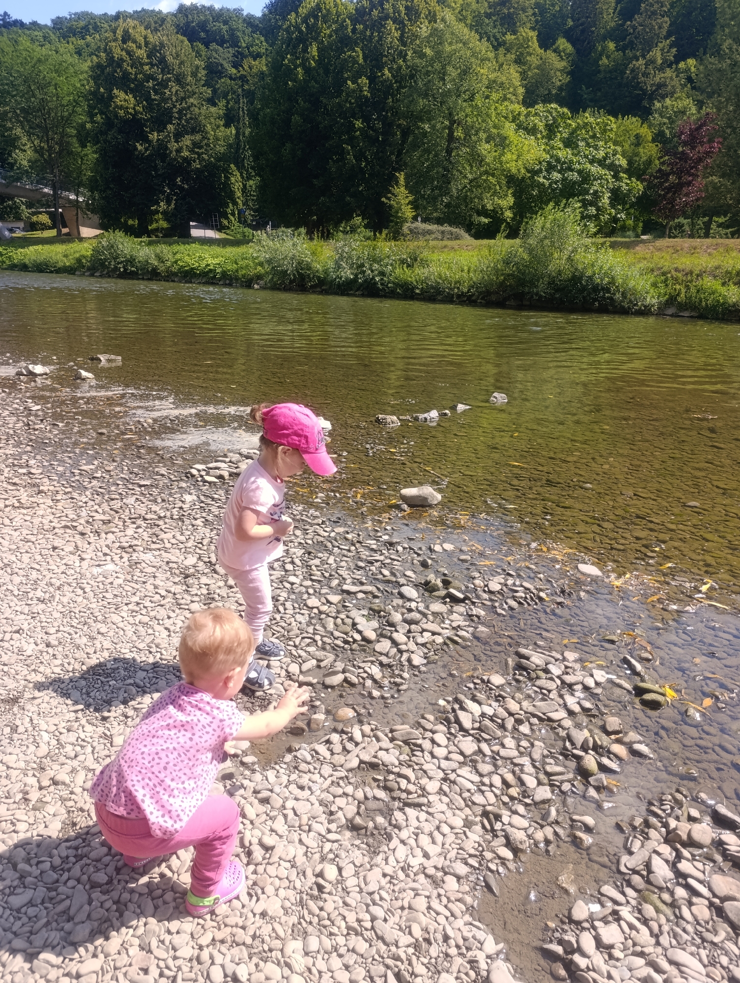 Two young children are playing on the rocky bank of a shallow, clear river, likely the Bečva River in Teplice nad Bečvou, Czechia. The child in the foreground, facing away from the camera, is crouched down and reaching towards the water. This child has blond hair and is wearing a pink t-shirt with a polka dot pattern and pink pants, with pink shoes. The second child, standing and facing left, is wearing a pink t-shirt, pink pants, and a pink baseball cap. Both children are dressed for warm weather. The riverbed is covered in smooth, grey and brown stones, with some small patches of mud visible where the water recedes. The water itself is clear, allowing the submerged stones to be seen, and has a greenish hue. The far bank of the river is densely wooded with mature green trees. The sky is a clear blue. The overall scene is one of outdoor recreation in a natural setting.