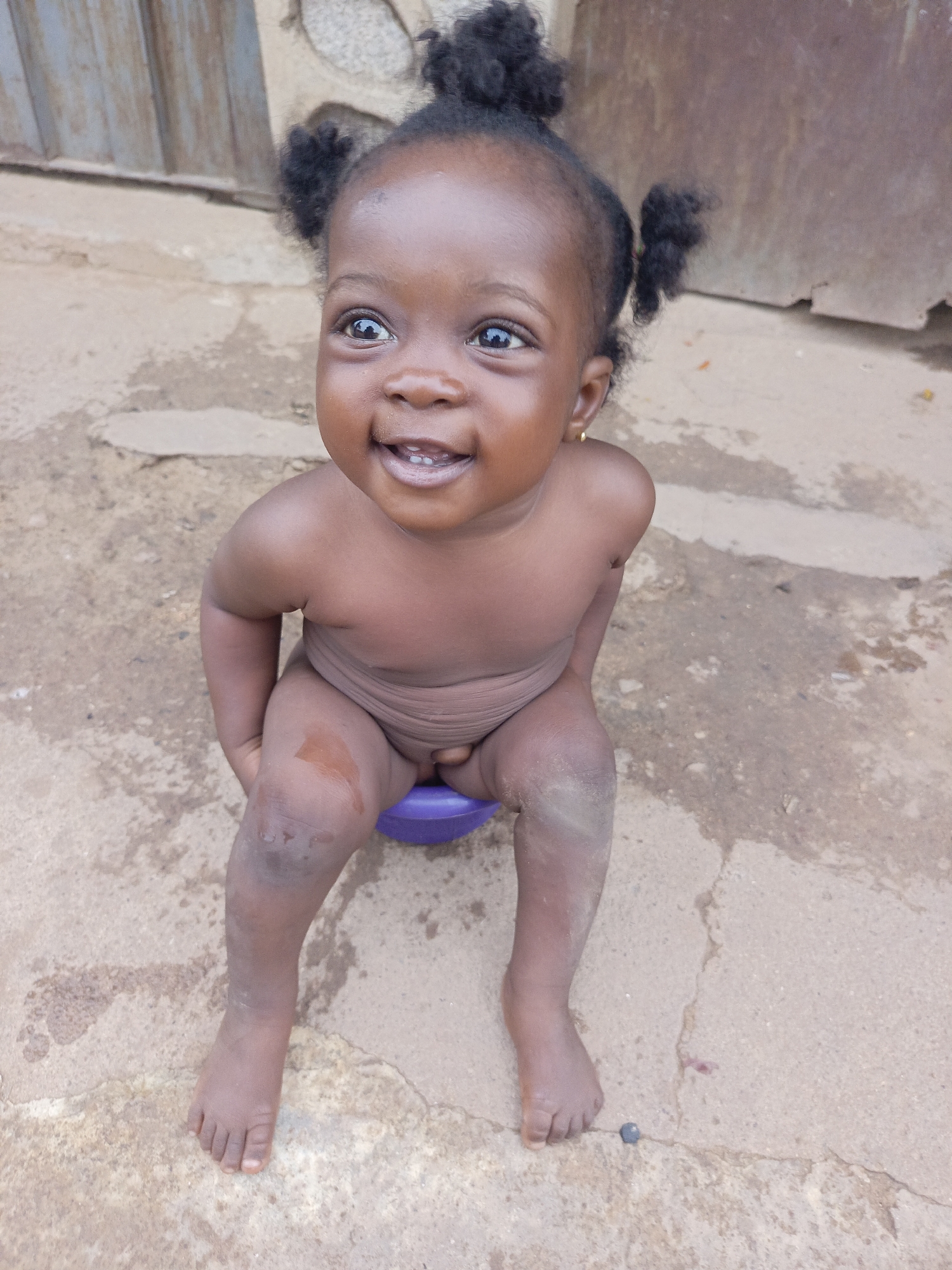 A toddler sits on a purple potty on a cracked concrete surface in Akwanga, Nigeria. The child has dark skin, two afro puffs on either side of their head, and is smiling, with a small gold earring visible. The child's body is unclothed and appears slightly wet or dirty, particularly on the knees. The background shows a metal door and a stone wall.