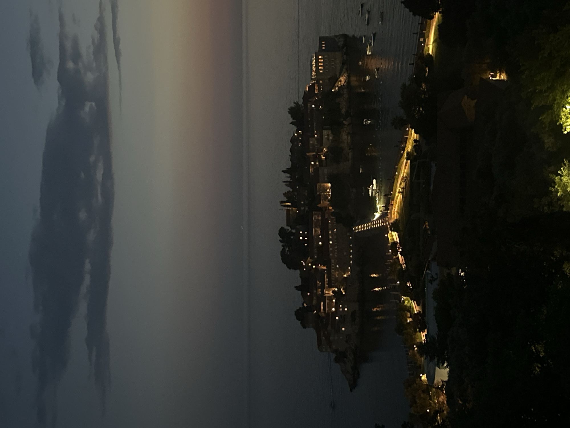 The image depicts a nighttime view of Sveti Stefan, a coastal town and island resort in Montenegro. The island, connected to the mainland by a narrow causeway, is illuminated by numerous lights from buildings and pathways. The sea surrounding the island is dark and calm, with a few small boats visible. The sky above is a deep twilight blue with scattered dark clouds. The foreground of the image shows a dark, wooded area, suggesting the viewpoint is from a higher elevation overlooking the town.