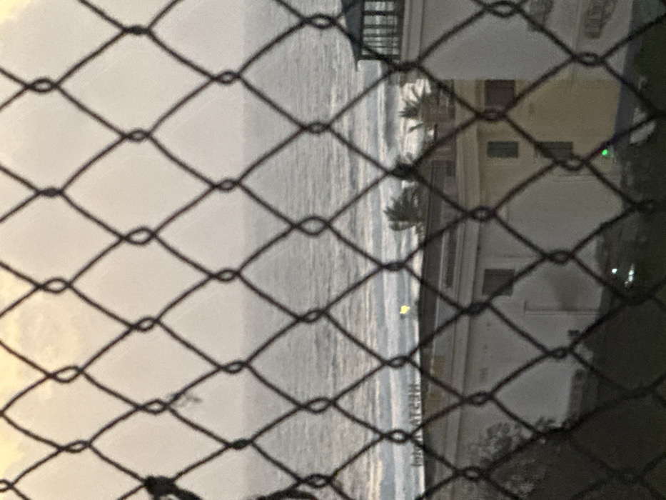 The image depicts a coastal view in Ngũ Hành Sơn Ward, Vietnam, observed through a blurred, dark chain-link fence that occupies the immediate foreground. The fence features diamond-shaped apertures. Beyond the fence, a body of water with choppy waves extends to the horizon under an overcast or hazy sky. Below the horizon, a narrow strip of coastline is visible. On the left side of the midground, a light-colored building is partially obscured by the fence, and what appears to be a reflective sign bearing the reversed text "HƎATƧƎЯ" (RESTAURANT) is visible near the waterline. A small, bright yellow light reflection is present on the water near this sign. Further to the right, a second light-colored building with dark windows is observed. To the far right, a cylindrical white structure with a dark, enclosed top, resembling a lighthouse or turret, stands prominent. Several palm trees are visible between the buildings and the ocean, with their fronds appearing to be affected by wind. Dark vegetation is visible in the lower left portion of the image. The ambient light suggests either dawn or dusk. No individuals are present, and no actions or interactions are depicted.