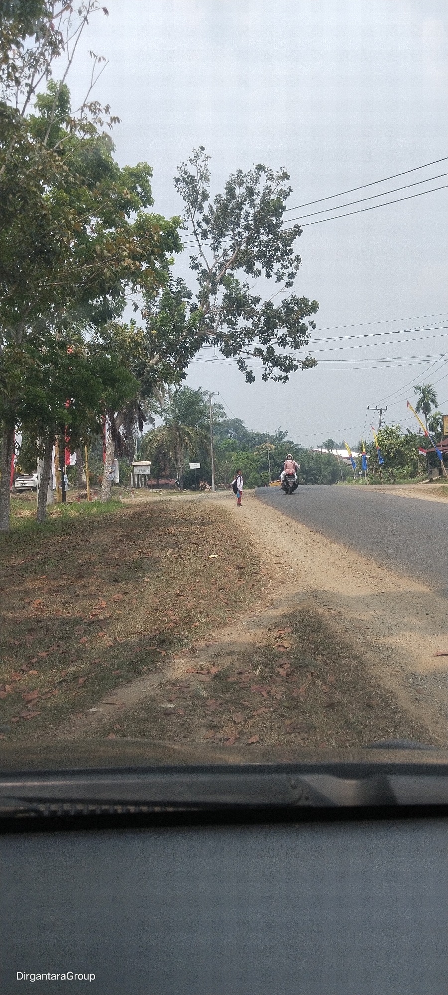The image depicts a roadside scene in Perintis, Indonesia, under an overcast sky. In the foreground, the hood of a vehicle is visible, with the "DirgantaraGroup" watermark in the lower left corner. A dry, grassy area with scattered fallen leaves lines the side of a paved road. A young person wearing a white shirt and blue shorts, carrying a backpack, stands on the side of the road facing away from the camera. Further down the road, a person is riding a scooter, also facing away. On the left side of the road, several trees are present, some adorned with red and white flags, possibly Indonesian flags. Beyond the road, dense green foliage and trees form a backdrop, with utility poles and wires stretching across the sky. The scene suggests a rural or semi-rural setting within Perintis.