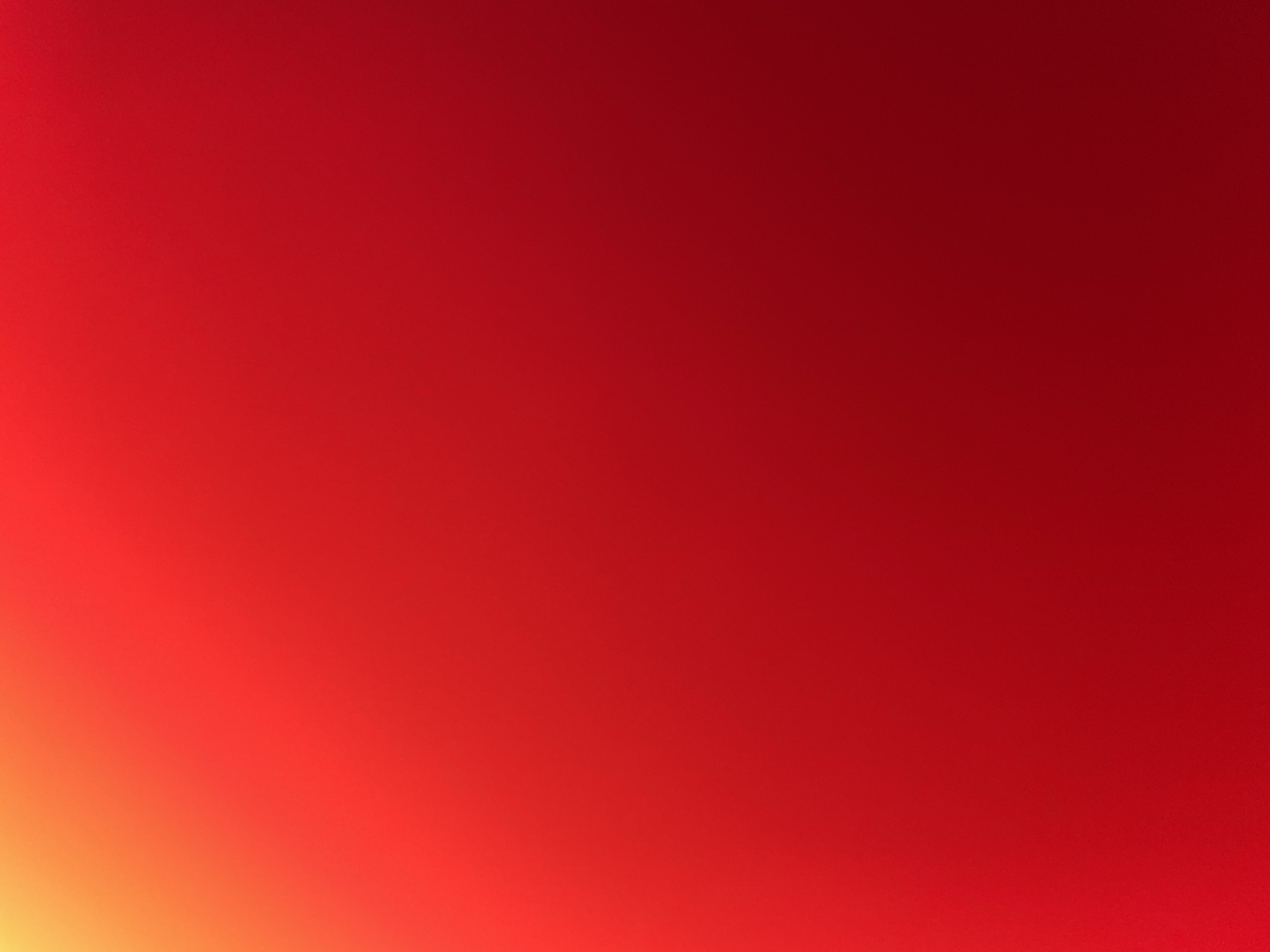 The image is a close-up, abstract shot of a red gradient. The color transitions from a brighter, yellowish-orange in the upper left corner to a deep, rich red across the rest of the frame. There are no discernible objects, people, or environmental features visible. The lighting suggests a soft glow or diffused light source, creating smooth color transitions.