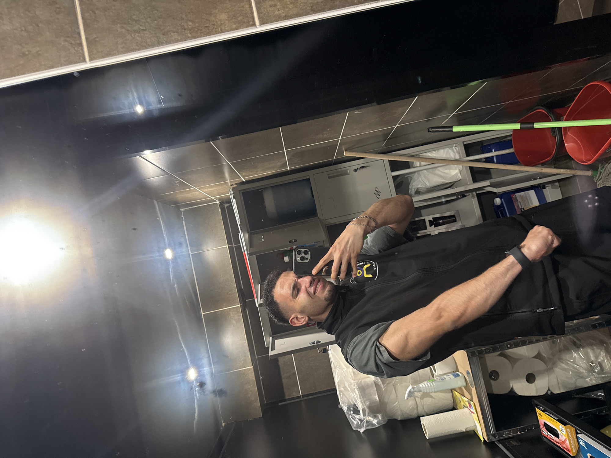 A man in a black vest is taking a selfie in a restroom or utility area. The area has grey tile walls and black countertops. A cleaning mop leans against the wall near two red buckets. Shelves hold various cleaning supplies, including toilet paper and bottled cleaners. A locker is situated behind the man. It is not possible to determine if the location is in Shumen, Bulgaria, based on the visual information provided.