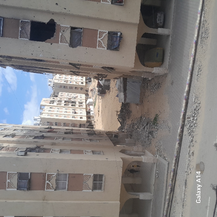 The image shows a street between buildings, appearing to be in a state of disrepair. Some windows are broken or covered. Debris litters the open space between the structures, suggesting damage or neglect. The buildings appear to be residential, but their condition implies a conflict zone or area of hardship.
