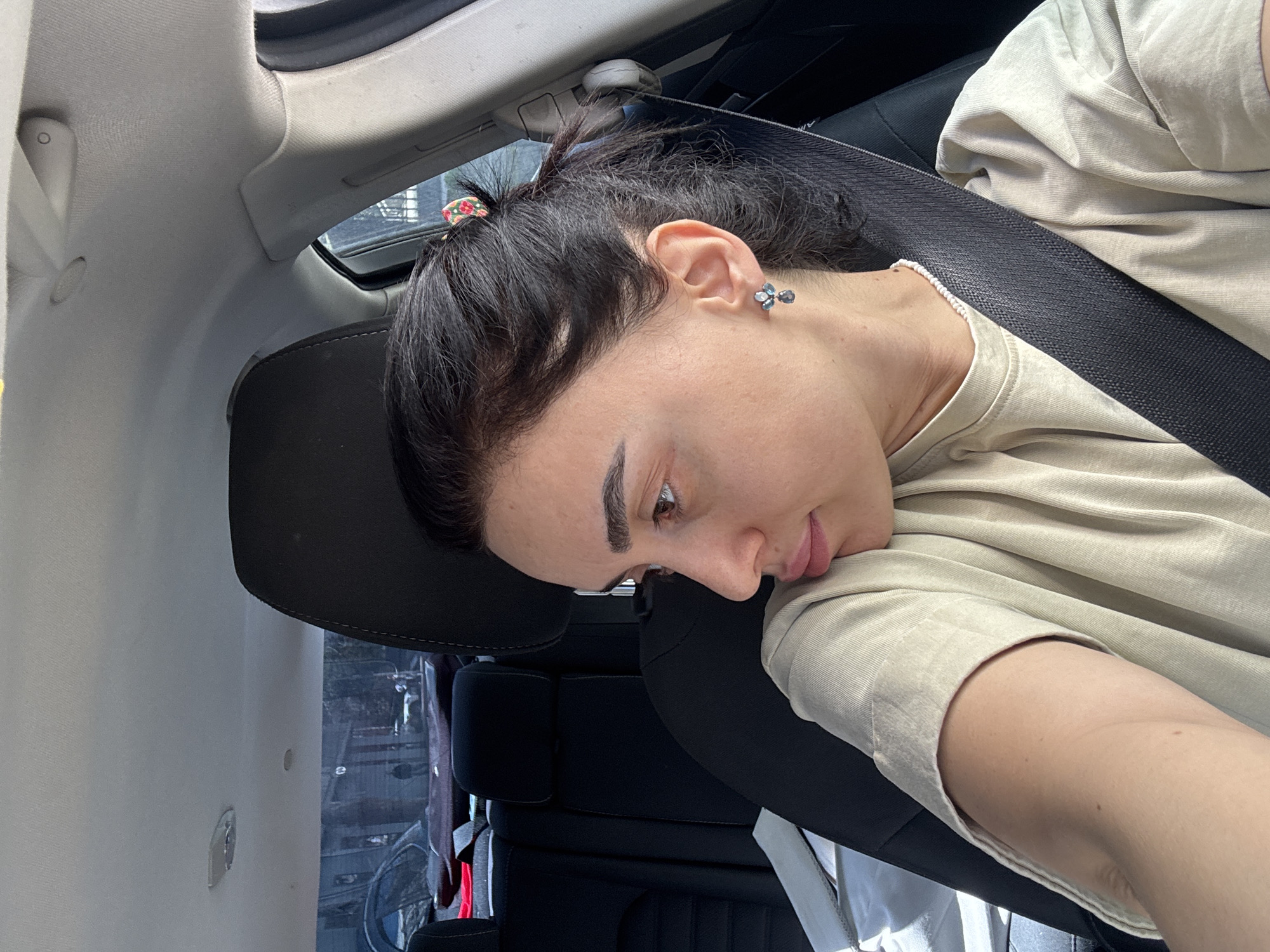 A young woman with dark hair pulled back in a small ponytail is sitting in the passenger seat of a car. She is wearing a light beige t-shirt and a black seatbelt. Her head is resting against the headrest of the seat. She has a stud earring in her left ear. The interior of the car is visible, including the dashboard and the window. The background outside the window suggests an urban environment, likely a street with buildings. There is no conclusive evidence of being in Tbilisi, Georgia.