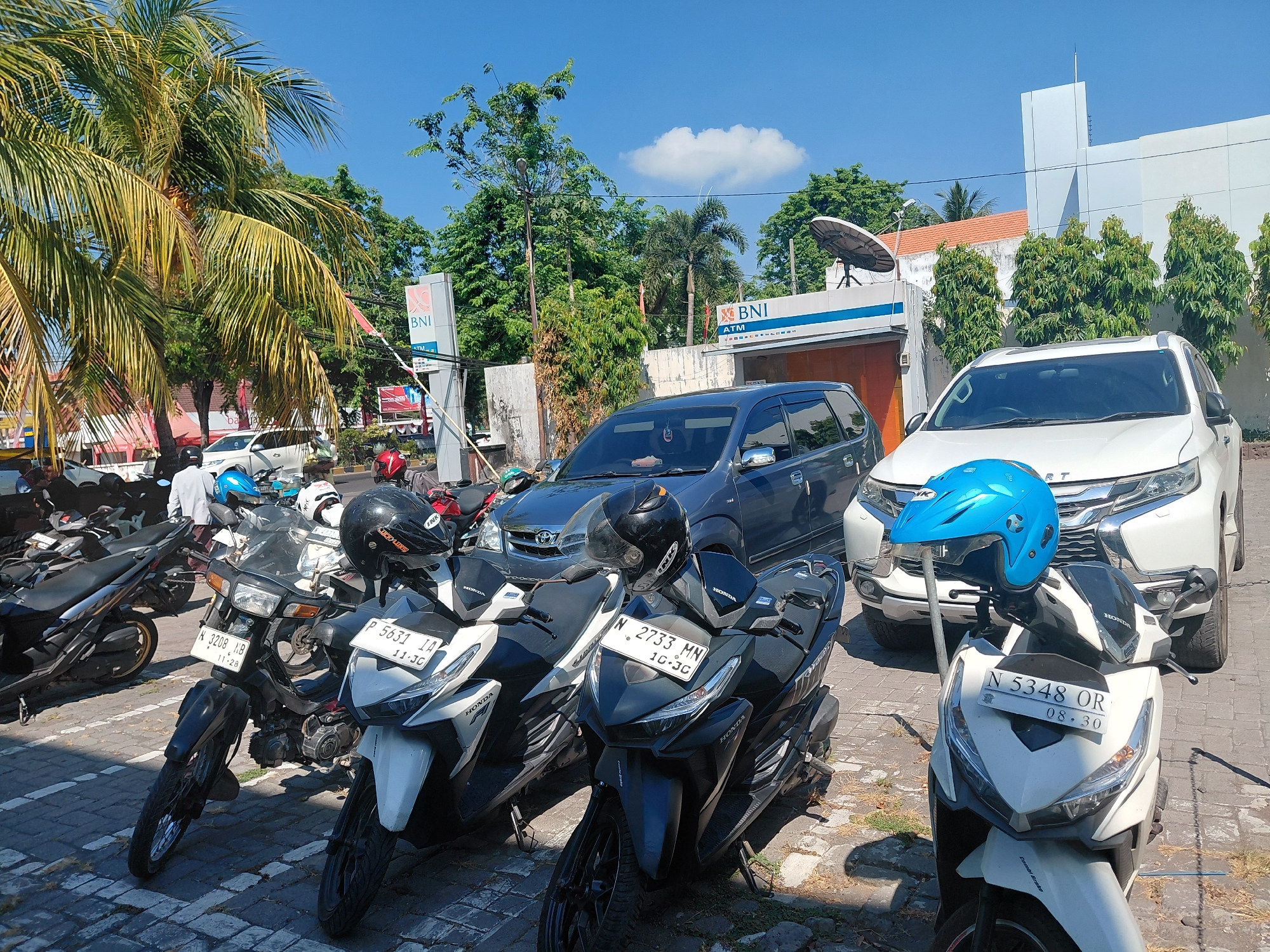 The image depicts a street scene in the city of Semampir, Indonesia, with a focus on parked motorcycles and vehicles. In the foreground, several scooters are parked on a paved surface. One white scooter in the lower right has a blue helmet placed on a metal pole in front of it. Another white scooter to its left has a black helmet. To the left of these, a dark-colored scooter is parked.
In the background, a dark blue minivan and a white SUV are parked. A building with the "BNI" logo, indicating Bank Negara Indonesia, is visible behind the vehicles. The building has "ATM" signage. Lush green trees and palm trees are present, suggesting a warm climate. The sky is clear blue with a few scattered clouds. The image appears to be taken during the daytime.