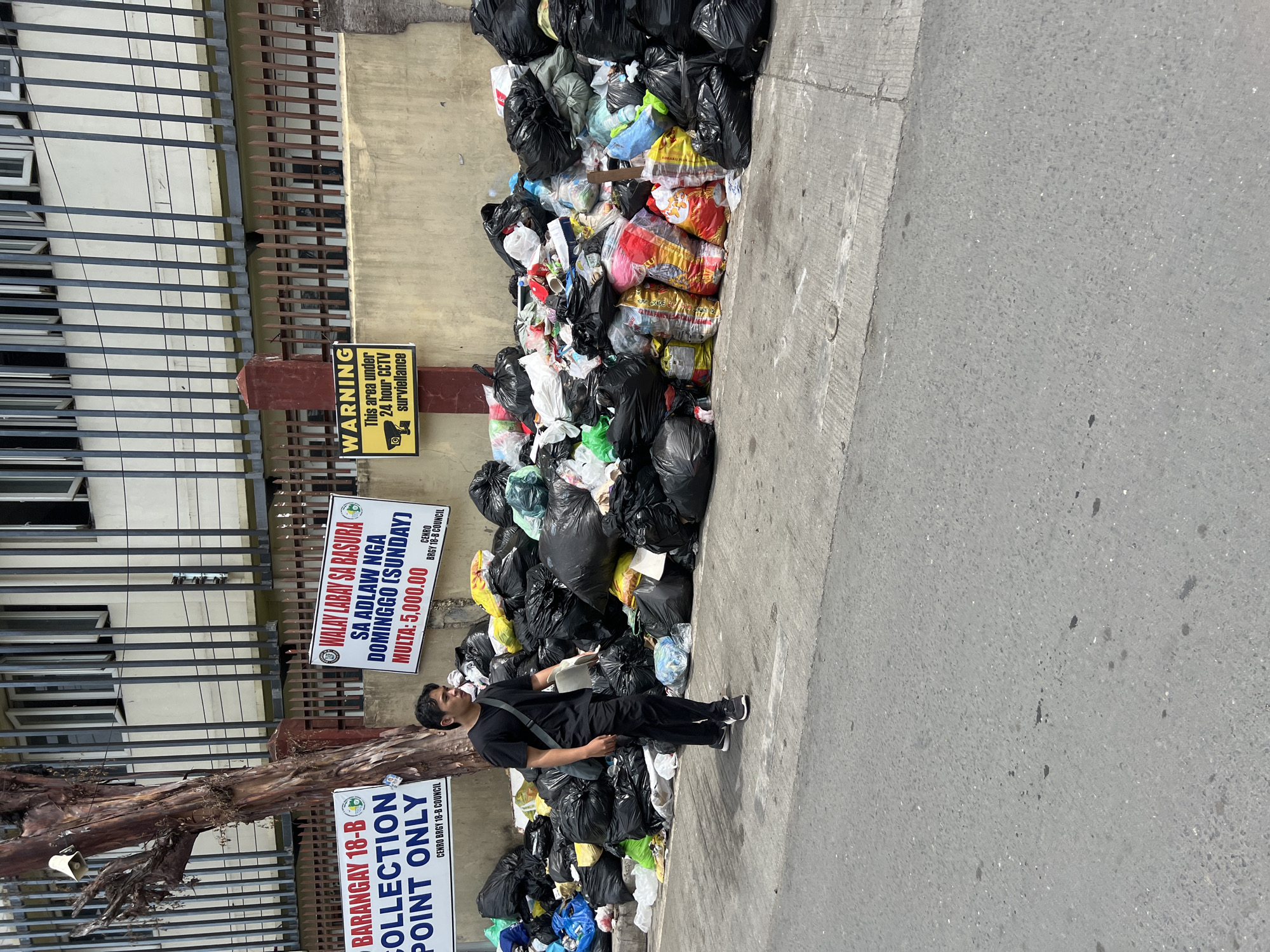 The image shows a large pile of garbage bags along a street in Barangay 18-B, Davao City, Philippines. The bags, mostly black, are stacked against a building and along the sidewalk. A man in a black shirt and pants stands near the garbage. Signs indicate the area is a collection point and dumping garbage on Sundays is subject to a fine. There is also a sign warning of 24-hour CCTV surveillance. The street appears to be concrete.