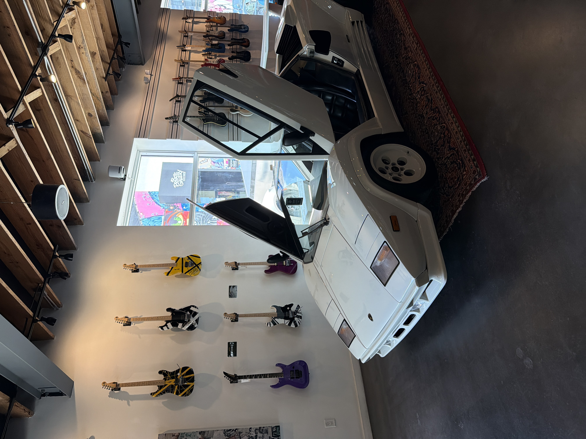 This image depicts the interior of a retail space, possibly a guitar shop or showroom, located in Miami, United States of America. A white, vintage Lamborghini Countach with its doors open is the central focus, positioned on a patterned rug. The car's interior, featuring black leather seats, is visible. Behind the car, an array of electric guitars are displayed on the wall, varying in color and design, including some with bold patterns. The background reveals a large window with graffiti art visible on the exterior, suggesting an urban setting. Industrial-style wooden beams and track lighting are present on the ceiling. The floor is a dark, polished concrete. No people are visible in the frame.