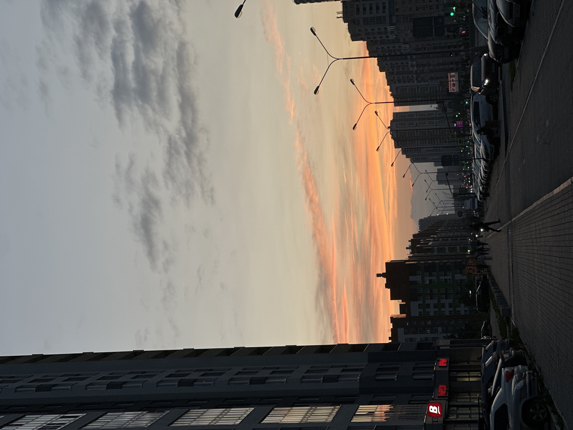 The image depicts a street scene in Yekaterinburg, Russia, at dusk. On the left side of the frame, a tall, modern apartment building dominates the foreground, its facade a dark gray with numerous windows reflecting the sky. The street stretches into the distance, lined with parked cars on the right and a sidewalk with pedestrians. Streetlights are visible along the road. The sky displays a gradient of colors, transitioning from a pale gray at the top to soft oranges and pinks near the horizon, indicating sunset. Buildings of varying heights and architectural styles line the street on the right, some appearing to be residential complexes with multiple floors and visible windows. Several individuals are walking on the sidewalk, some in silhouette against the fading light. The overall atmosphere is calm, suggesting the end of the day in an urban environment.