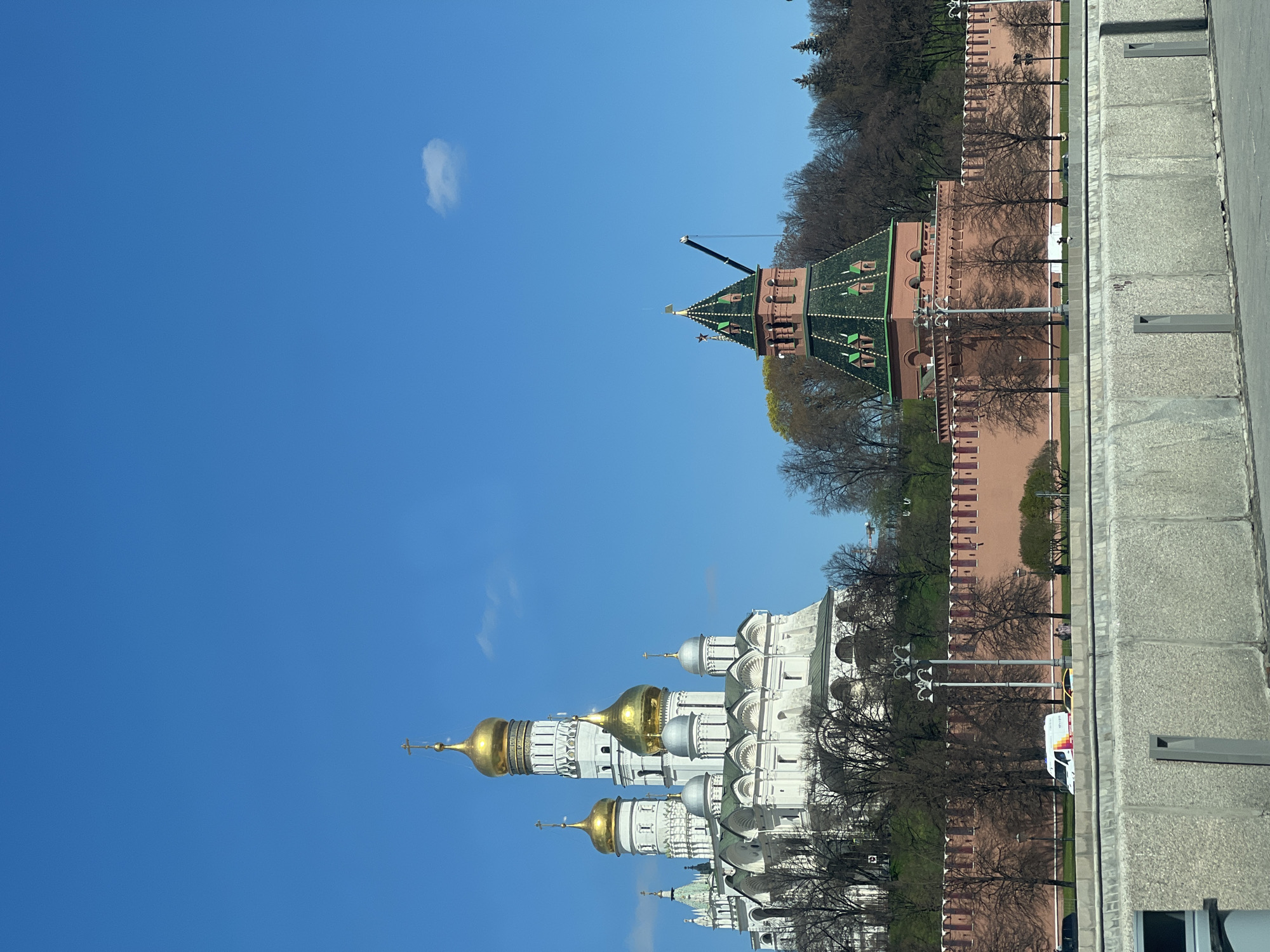 This image showcases the Moscow Kremlin, a fortified complex in Moscow, Russia, overlooking the Moskva River to the south, Saint Basil's Cathedral and Red Square to the east, and the Alexander Garden to the west. The frame includes a view of the Kremlin's walls and one of its towers, along with a collection of white-stone cathedrals topped with golden domes. The scene is set on a clear day with a bright blue sky. A stretch of pavement and a wall run along the bottom-right edge of the frame. The photograph's orientation is sideways, with the sky occupying the majority of the left side and the Kremlin structures and trees positioned on the right.