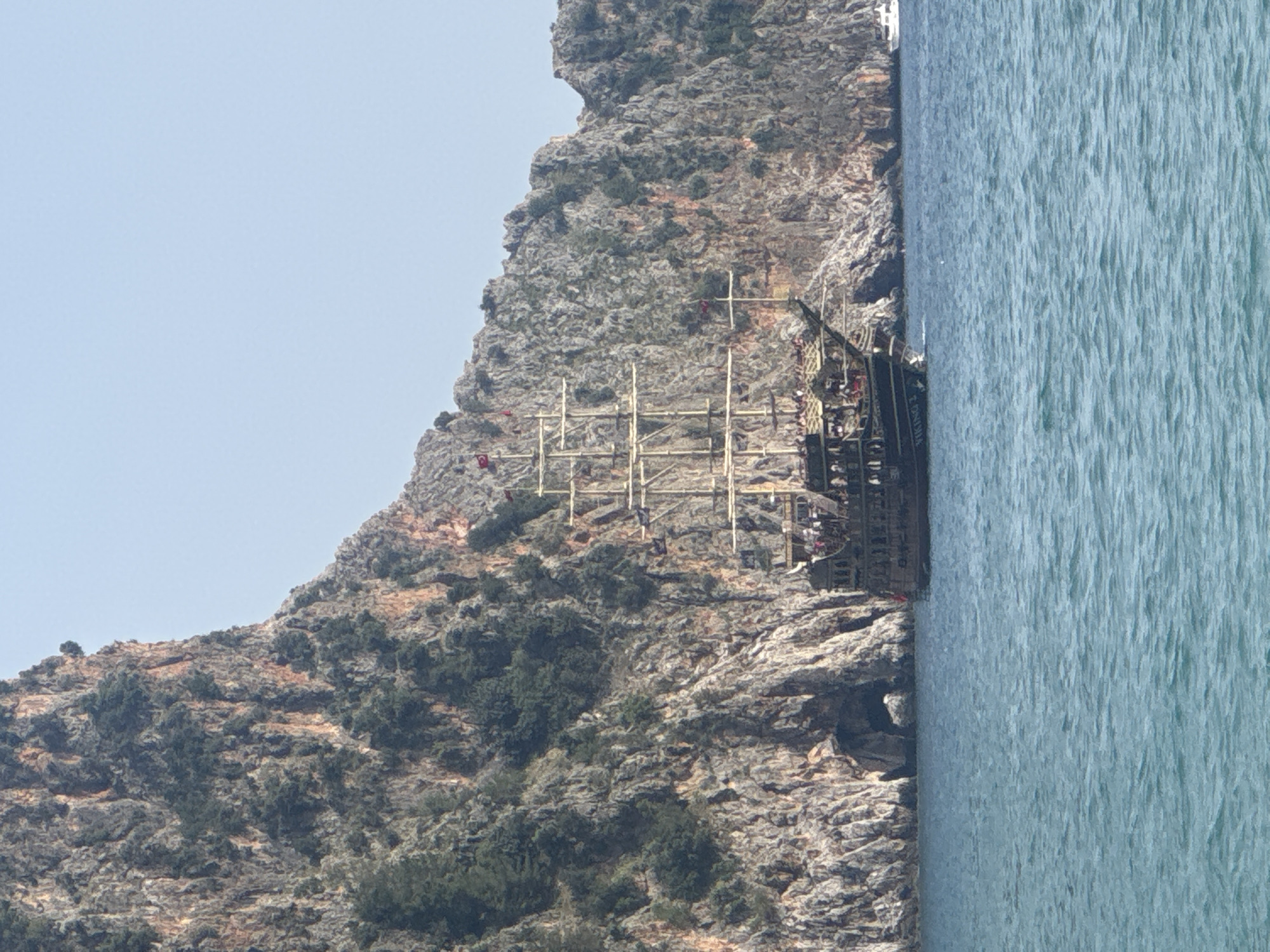 The image shows a coastal scene in Alanya, Turkey. A large, ornate tourist boat modeled after a pirate ship is visible in the water, docked along a rocky cliffside. The ship has multiple masts and is crowded with people. The water appears calm. A rocky mountain rises from the coastline. The sky is clear and light blue.
