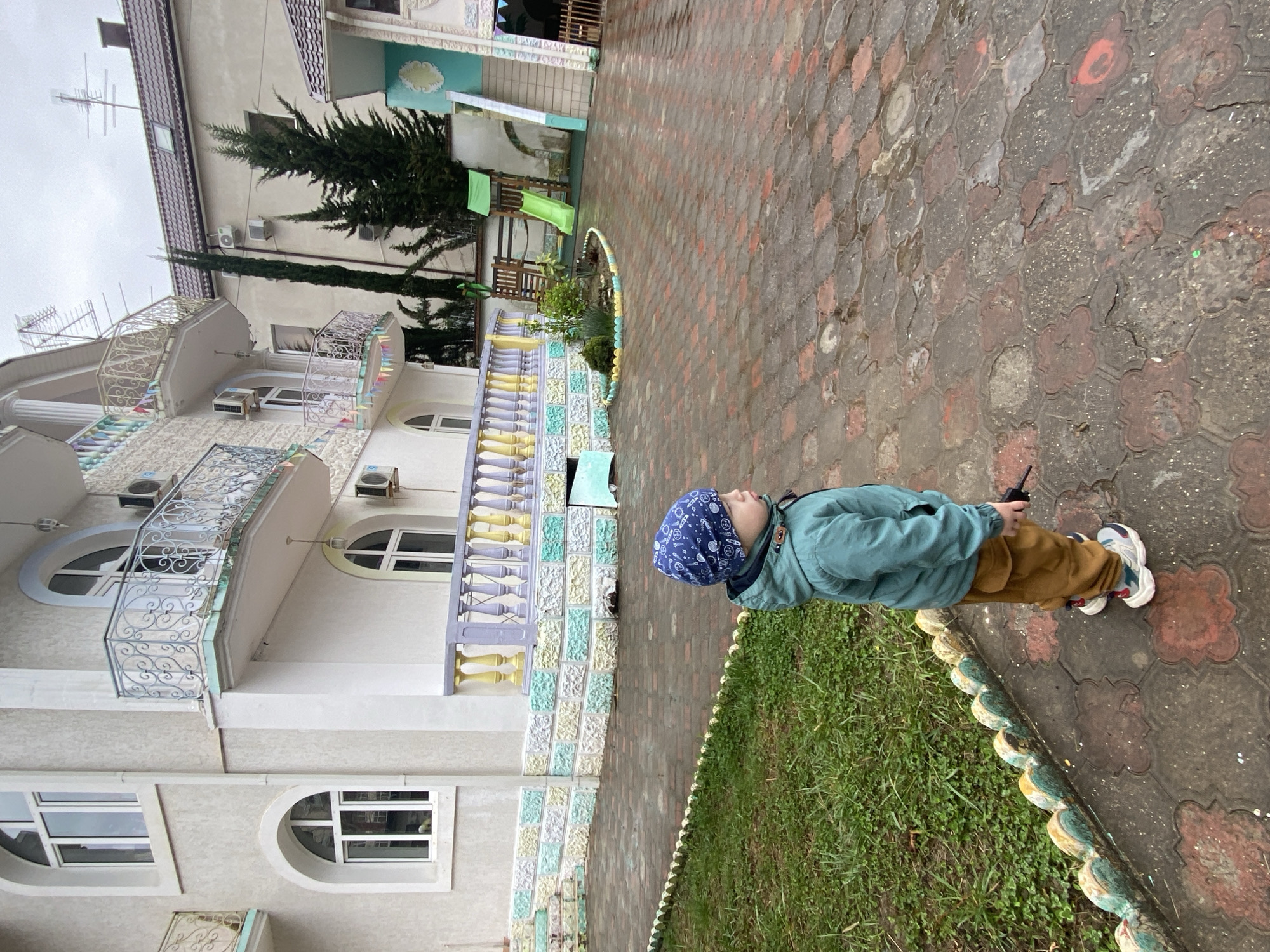 A young boy wearing a blue hat, a light green jacket, and brown pants stands on a patterned red and gray brick walkway. He is holding a black object, possibly a walkie-talkie. To his left is a building with white walls, arched windows, and decorative railings. A border of green grass and decorative stones runs along the edge of the walkway, separating it from the building. The location appears to be outdoors, potentially in a residential area or park in Sochi, Russia.