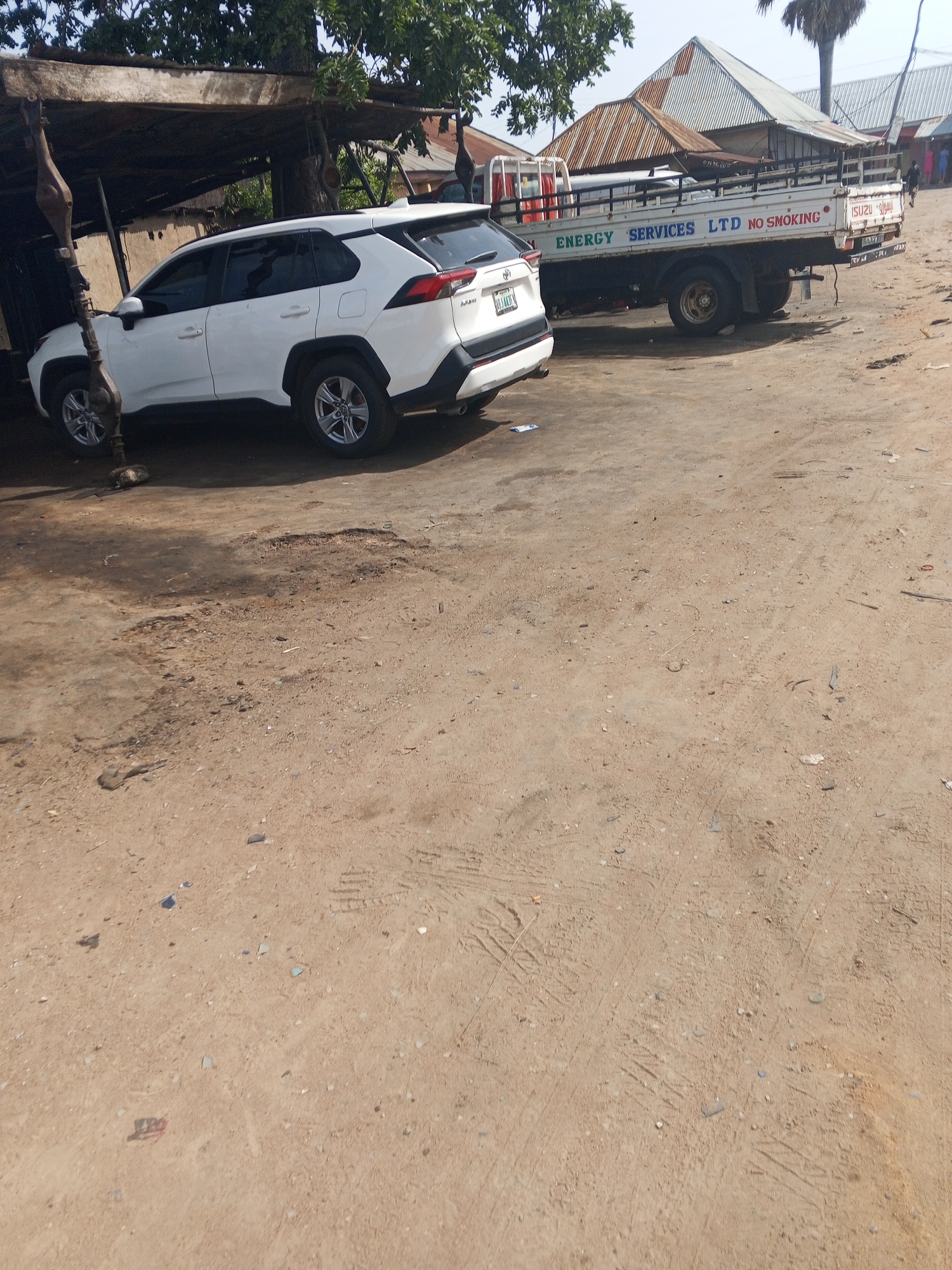 The image depicts a street scene in Jalingo, Nigeria, with two vehicles parked on a dirt and cracked asphalt surface. On the left, a white Toyota RAV4 SUV is parked under a weathered, makeshift shelter with a corrugated metal roof. To the right of the SUV, a flatbed truck with the branding "ENERGY SERVICES LTD NO SMOKING" and "ISUZU" is also parked. The truck has a white cab and a light-colored bed with a metal railing. In the background, more buildings with corrugated metal roofs are visible, suggesting a commercial or industrial area. The ground is uneven, with patches of loose dirt and visible tire tracks. The overall environment appears dusty and somewhat dilapidated. No people are clearly visible in the immediate vicinity of the vehicles. The lighting suggests it is daytime.
