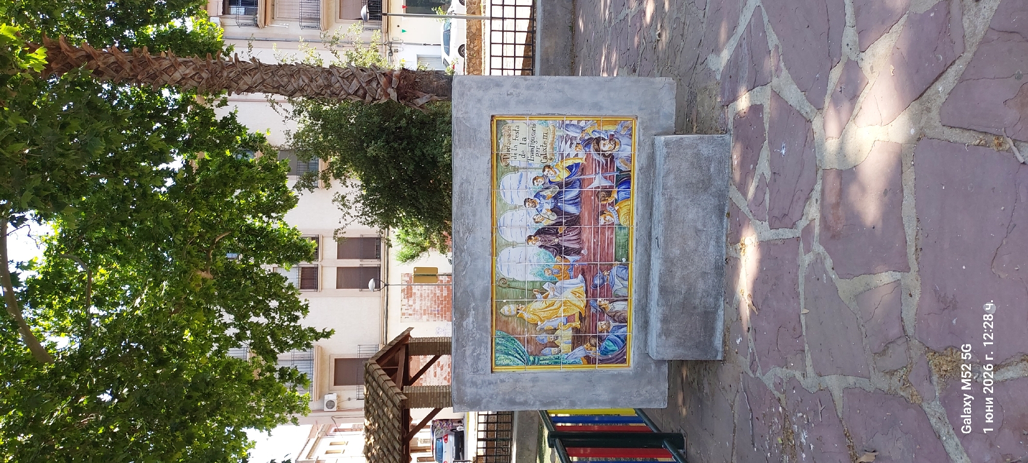 Here is a detailed description of the image:
The image shows a tiled monument in what is presumed to be a public area, likely a park or square, in the city of Canals, Spain. The monument features a large tile mural depicting a historical scene, possibly related to the "Proclamacio de la festa de la Transfiguracio per Calixte III". The mural shows figures in historical attire.
The monument is made of concrete with a rough, weathered texture. It is situated on a stone-paved surface. Various plants, including trees, shrubs, and a palm tree, are visible in the background and to the sides of the monument. Parts of a building are also visible behind the monument. A metal fence and what appears to be a set of colorful benches are also partially visible.
There is some text on the image indicating it was taken with a Galaxy M52 5G phone on June 1, 2026, at 12:28.