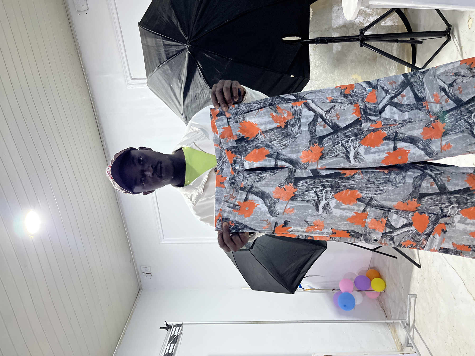 The image depicts a male individual standing indoors, holding up a pair of camouflage pants. The person has dark skin, a mustache, and is wearing a light-colored jacket over a bright green shirt, topped with a multi-colored baseball cap. They are centered in the frame, holding the pants horizontally across their chest. The pants feature a gray, black, and white tree bark pattern with prominent bright orange maple leaf designs and cargo pockets.
The setting appears to be a photography studio or similar space with white walls and a white paneled ceiling. A bright, circular overhead light fixture is visible on the ceiling. Two large black photographic umbrella softboxes are present; one to the left and one to the right of the individual, with the right one standing on a black tripod light stand. A metallic rack or stand is positioned on the far left. In the mid-left background, a cluster of colorful balloons (blue, pink, purple, yellow, orange) rests on the floor. A partial view of a white door frame is visible on the right wall, and a small, round white table or stool is partially visible on the far right. The floor is light-colored. The location is Unknown, Unknown.