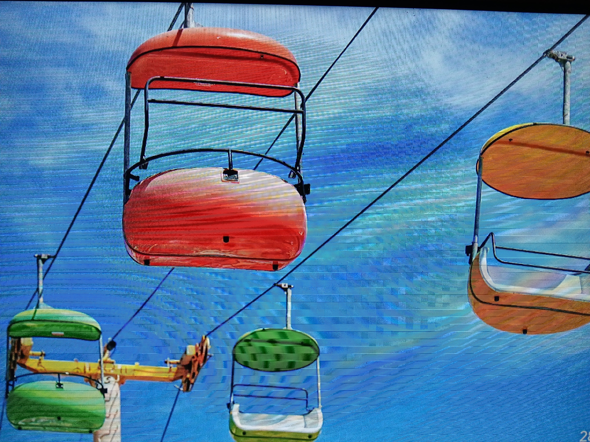 The image shows a cable car system with several gondolas of different colors against a blue sky. One set is orange, another green, and another yellow. The city of Juan Griego, Venezuela, is not readily apparent in the image. The scene is focused on the cable car infrastructure and the gondolas.