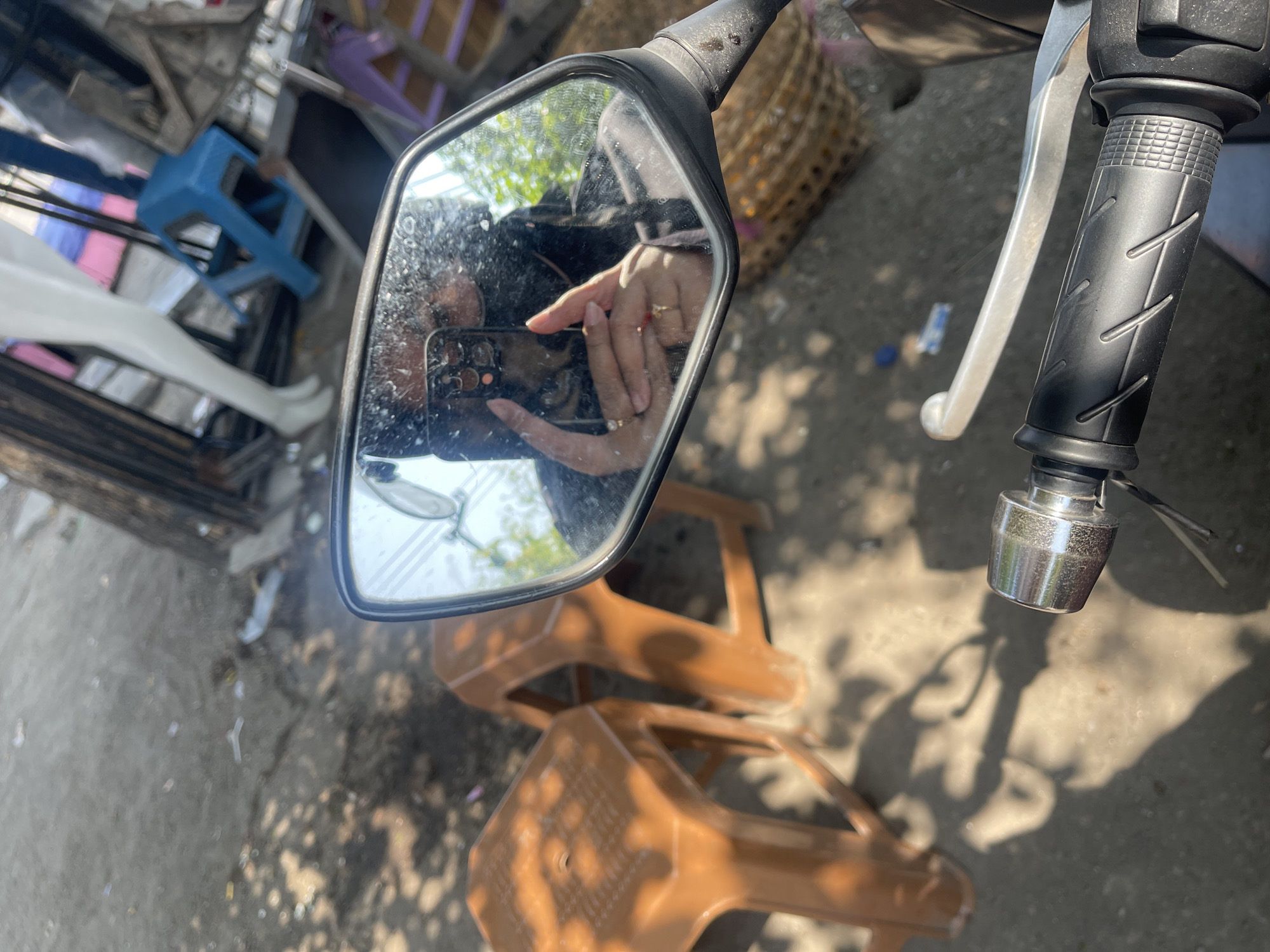 A mirror on a motorcycle reflects a person taking a selfie. The person is wearing a dark hijab and a dark jacket, and is holding a smartphone in their left hand. They are also wearing two rings on their left hand. In the background, there are two brown plastic stools, a basket, and some debris on the ground. The scene appears to be outdoors, possibly in a marketplace or a roadside area in Balongdowo, Indonesia, given the presence of a mannequin and other items commonly found in such locations. The lighting suggests it is daytime, with shadows cast by overhead foliage.