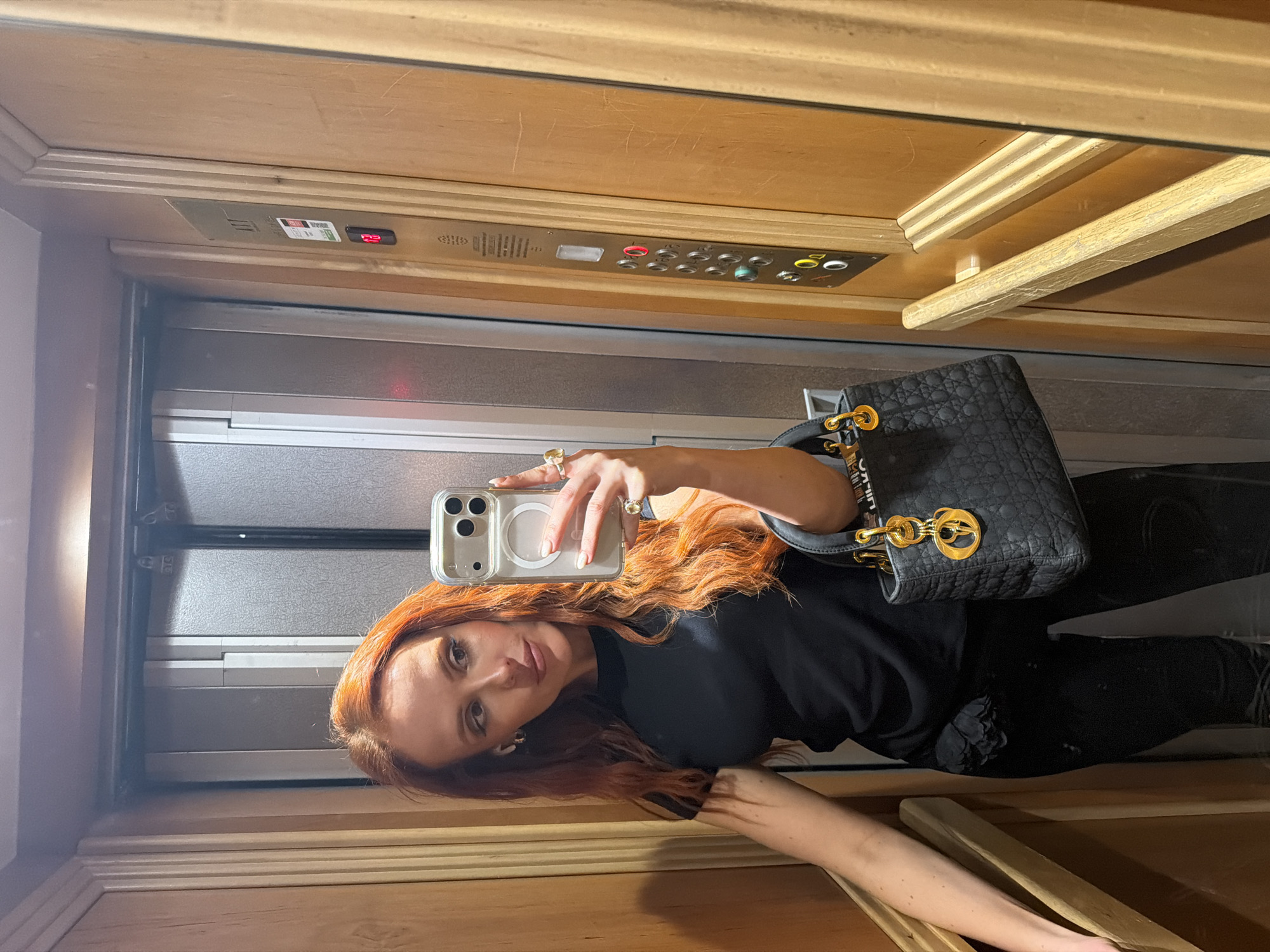 A person with red hair is taking a selfie in an elevator. The elevator has wooden interior walls and a control panel with buttons and a floor display showing the number "23". The person is holding a smartphone and wearing a black outfit with a black handbag. Based on the style of the elevator and potential for luxury fashion, it is possibly located in Paris, France.