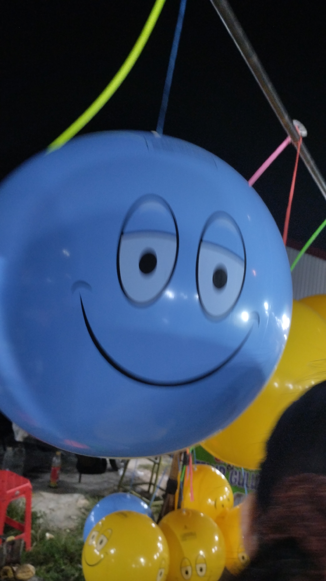 A large, light blue balloon with a cartoon face, featuring wide, black-pupiled eyes and a wide smiling mouth, dominates the foreground of this outdoor image. The balloon is suspended by colorful strings, including lime green, blue, and pink, which are attached to a metal pole overhead. The scene appears to be an outdoor fair or event, likely at night or dusk, given the dark background and the bright artificial lighting reflecting off the balloons.
Behind the main blue balloon, several yellow balloons are visible, some also decorated with cartoon faces, suggesting a festive atmosphere. One yellow balloon in the lower right has a furry, dark object partially obscuring it, possibly a person's clothing or hair. In the lower left, a red stool and some greenery can be seen, indicating a ground-level setting. The overall impression is one of a cheerful and playful gathering. No visible text is present in the image. The location context of Sambungrejo, Indonesia, suggests this could be a local celebration or market.