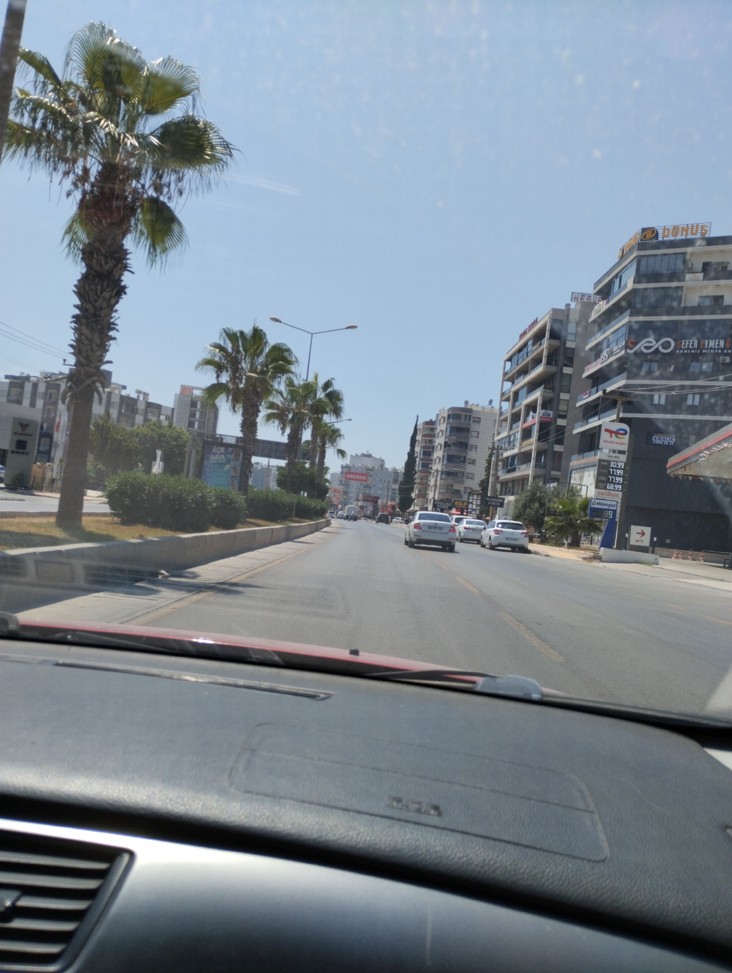 The image is a first-person perspective from inside a vehicle, looking forward through the windshield at a city street in Mezitli, Turkey. The street is paved and has traffic lanes marked by yellow lines. Several white cars are driving on the road, some in the foreground and others further in the distance.
On the right side of the street, there are several buildings. The closest building is a modern, multi-story structure with a dark facade, featuring several floors with windows. A prominent sign on this building reads "TotalEnergies" with fuel prices displayed below. Further down the street, other buildings of varying heights and architectural styles are visible.
On the left side of the street, there are palm trees, some of which are tall and mature, lining the sidewalk. There are also smaller trees and green bushes. A concrete barrier separates the sidewalk from the road. In the background, more buildings and streetlights can be seen. The sky is clear and blue, indicating a sunny day. The overall impression is that of a typical urban scene on a bright day.