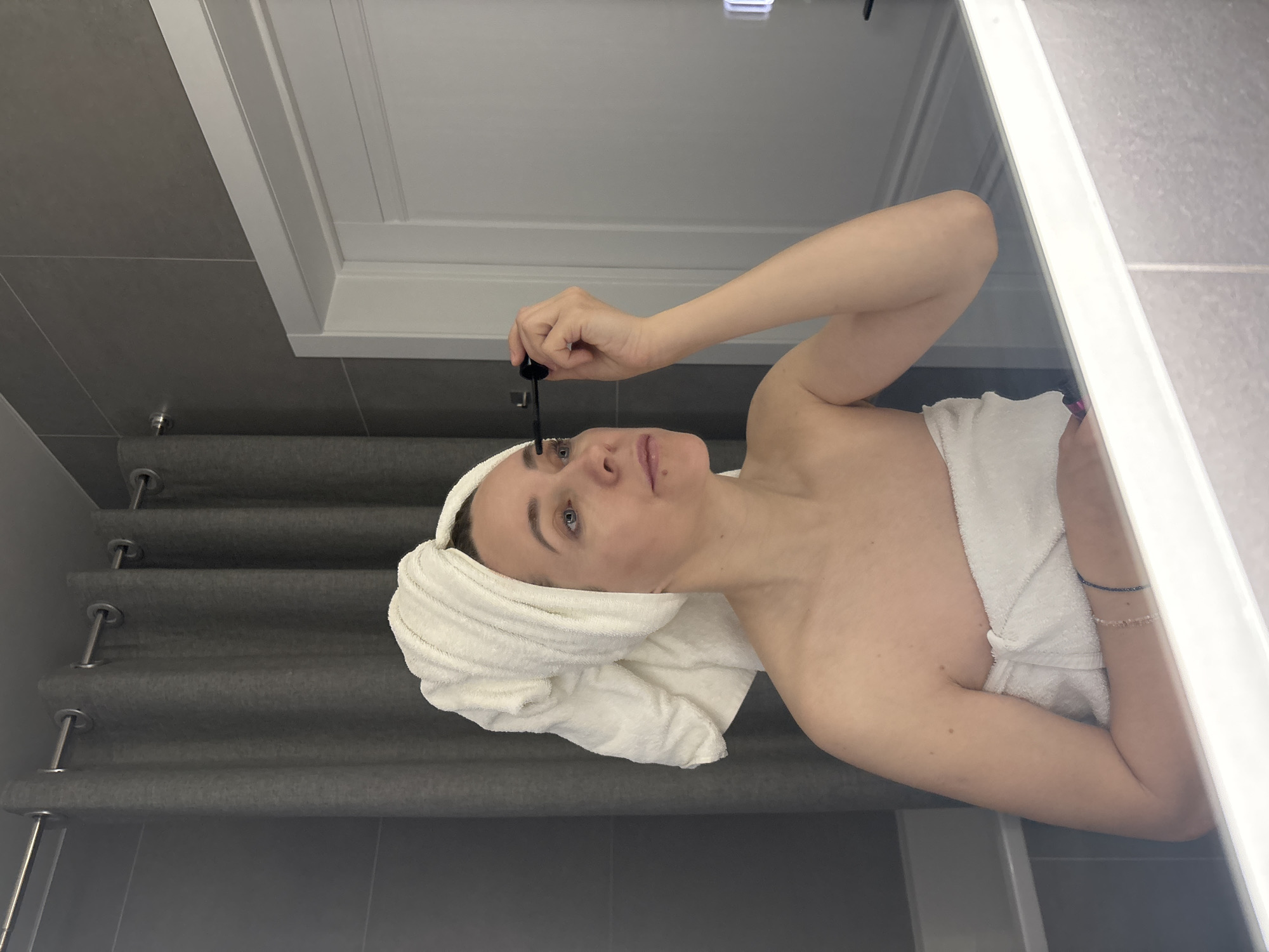 A woman with fair skin is applying an unknown liquid to her eyelashes in a bathroom. She has a white towel wrapped around her head and body. She holds a black applicator in her right hand, bringing it close to her right eye. The bathroom has gray walls, a white door, and a metal curtain rod and curtain. The location in the city of Chaikovsky, Russia, is not discernible from the visual information.