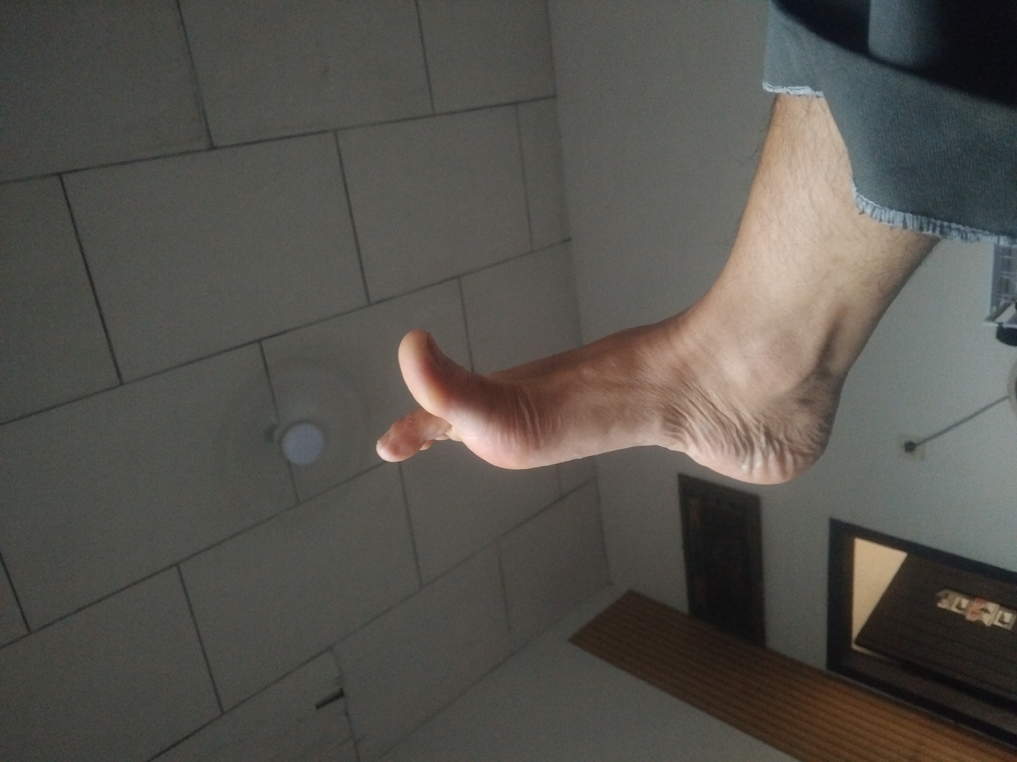 The image shows a close-up view of a person's bare foot, positioned from the ankle to the toes, with the sole facing upwards. The foot is resting on a grey surface, possibly a bed or couch, with a glimpse of dark grey fabric with a frayed edge visible on the right side. The background consists of a white ceiling with a grid of square tiles, and a faint circular light fixture can be seen. To the left of the foot, a section of a wooden wall paneling and a framed picture on the wall are partially visible. A doorway with some text on it is also visible in the lower left corner. There is no indication of the city of Tempeh, Indonesia, within the image itself; it is an unrelated piece of information.