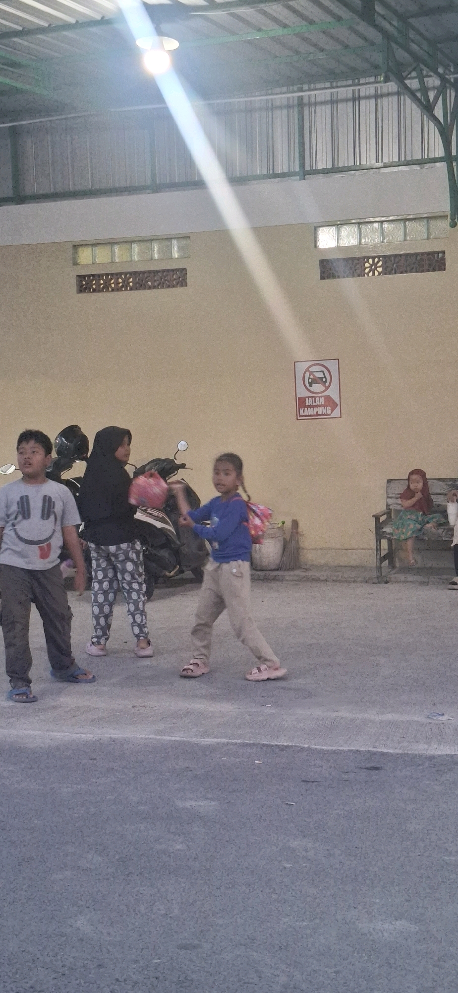 In Desa Ngesrep, Indonesia, the image captures an outdoor scene under a covered structure with a metal roof and green supports. The ground is paved with asphalt.
In the foreground, three children are visible. On the left, a young boy wearing a grey t-shirt with a smiley face and dark pants stands with his mouth slightly open, looking upwards. Next to him, a person wearing a black hijab and patterned pants is positioned behind a motorcycle. To the right of the person in the hijab, a young girl in a blue long-sleeved shirt and beige pants is actively gesturing with her hands, appearing to be in motion, possibly playing or interacting with someone out of frame. She carries a pink backpack.
Behind the children, a motorcycle is parked. The wall of the building is a light beige color. On the wall, a sign with a red circle and a diagonal line across a vehicle symbol, accompanied by text that reads "JALAN KAMPUNG" (Village Road), is displayed. To the far right, a young girl wearing a red headscarf and a patterned dress sits on a wooden bench.
The lighting suggests it might be late afternoon or early evening, with a bright light source casting a strong diagonal beam across the scene from the upper left.