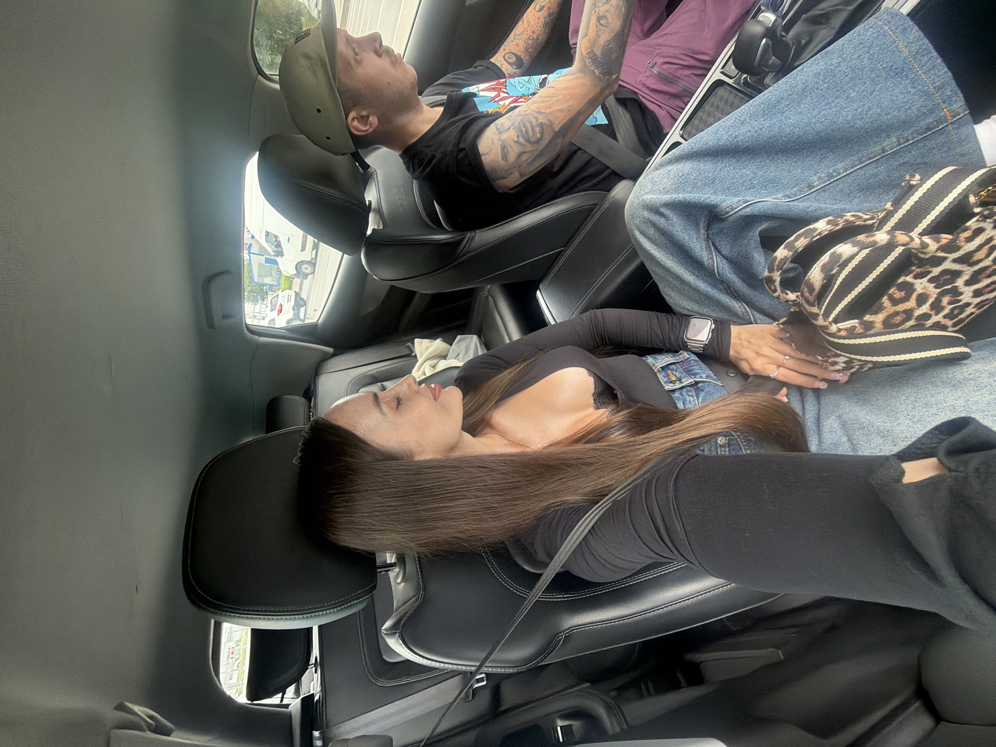 In the backseat of a car, a woman with long brown hair and closed eyes sits in the left seat. She wears a black long-sleeved shirt, a black bra visible at the neckline, and wide-leg denim jeans. A leopard-print and striped bag rests on her lap, and a silver smartwatch is on her left wrist. To her right, a man with a green baseball cap and multiple tattoos on his arms wears a black t-shirt and purple pants. He is looking forward, presumably driving. The car's interior is visible, including black leather seats and a center console. Outside the car windows, other vehicles are on a city street, indicating an urban environment. The lighting suggests it is daytime. The scene is likely Saint Petersburg, Russia, given the context of the request.