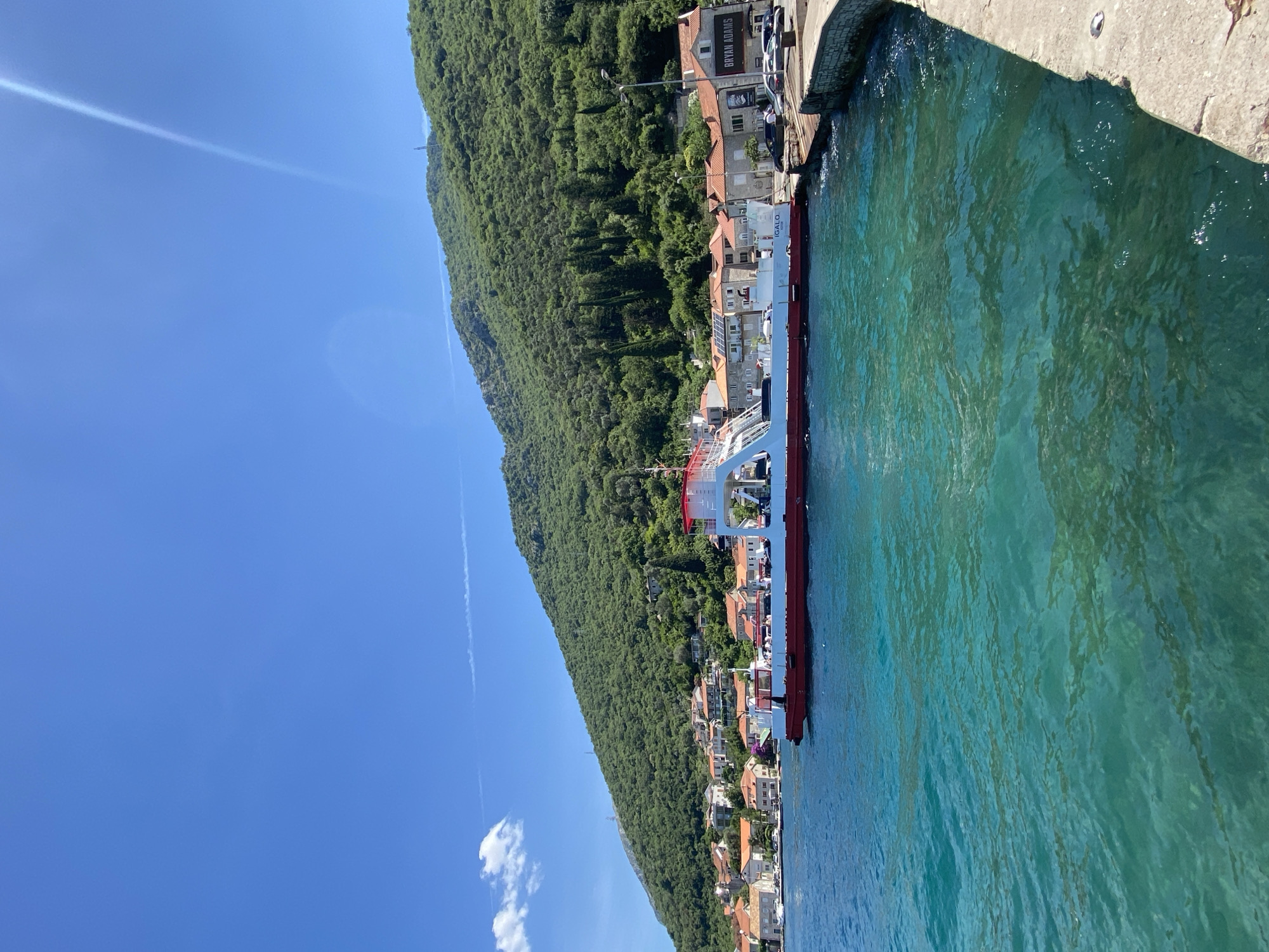 A ferryboat with red and white coloring is docked at a pier in Lepetane, Montenegro. The ferryboat is positioned in the foreground, partially obscuring a line of buildings with orange-tiled roofs. Behind the buildings, a lush green mountain slopes upwards, with scattered trees and a clear blue sky above. The water in front of the ferryboat is a vibrant turquoise, with visible ripples and some darker patches indicating shadows or submerged objects. The pier is made of concrete and extends into the water from the right side of the frame. A few cars are parked along the waterfront. The overall scene depicts a sunny day in a coastal town.