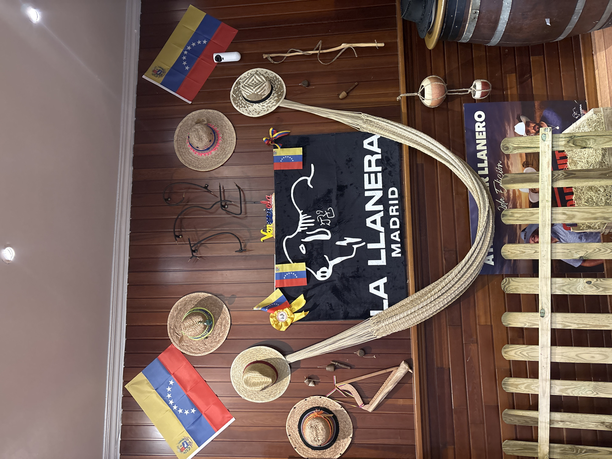 The image depicts the interior of a Venezuelan restaurant, "La Llanera," located in Torrejón de Ardoz, Spain. The walls are adorned with Venezuelan flags, traditional straw hats, and decorative items like a hammock, a small harp, and various trinkets. A prominent black banner displays the restaurant's name and location, "LA LLANERA MADRID," along with a stylized outline of a cow. In the foreground, a rustic wooden fence encloses a display of hay, and a poster for "La Llanera Edicion" features a man in a cowboy hat. To the right, a large wooden barrel and a traditional Venezuelan hat contribute to the cultural ambiance. The lighting suggests an indoor setting, possibly a dining area or a themed section of the restaurant.