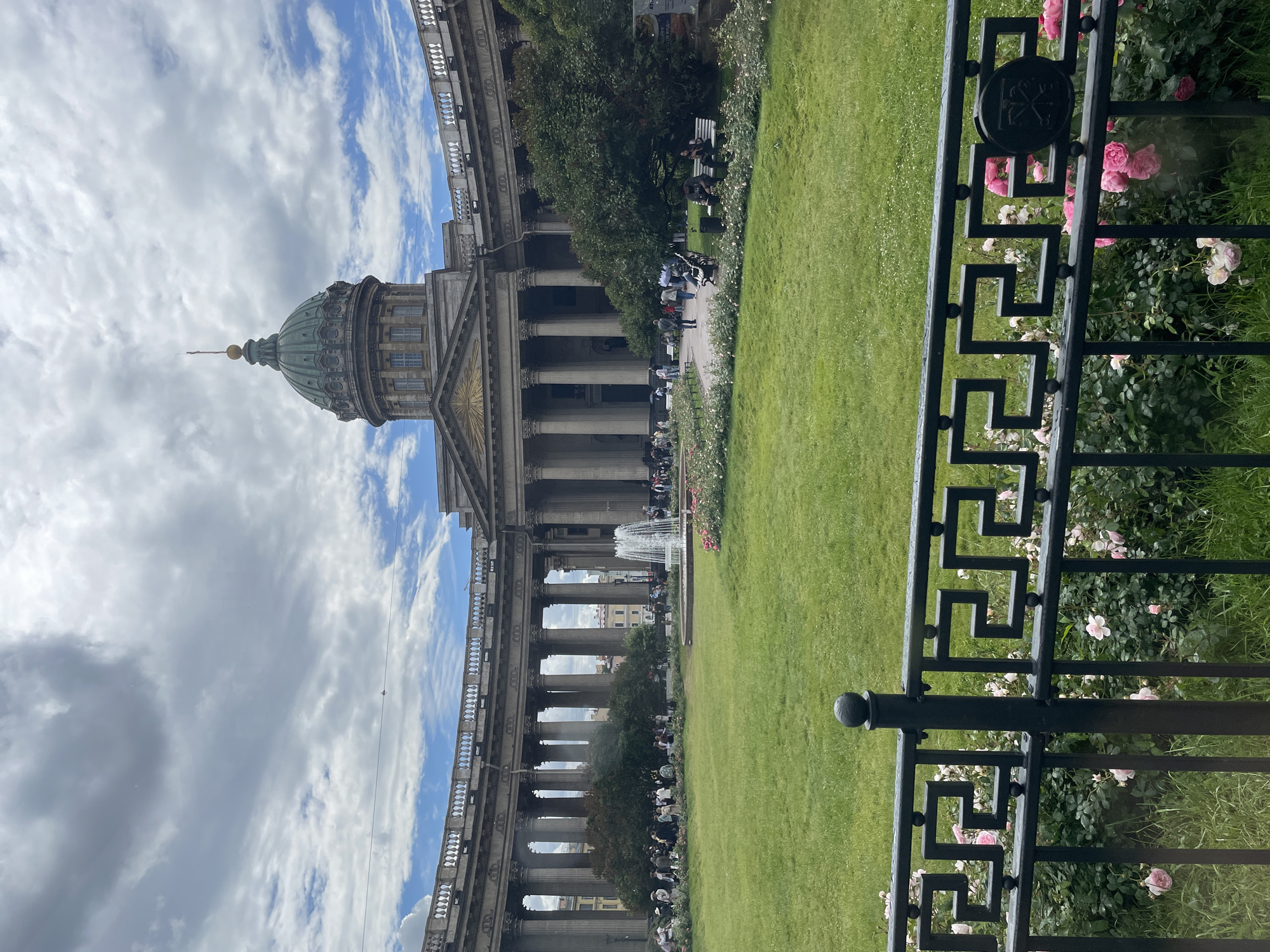 The image shows the Kazan Cathedral in Saint Petersburg, Russia. The grand neoclassical building features a large colonnade and a green dome. In the foreground, a black ornate metal fence separates the viewer from a lush green lawn with scattered pink roses and bushes. Several people are visible in the middle ground, some sitting on benches, others walking along a path. A fountain is also present in the middle ground, with water jets spraying upwards. The sky is partly cloudy with a mix of blue sky and white clouds. The overall scene suggests a pleasant day, possibly late morning or early afternoon, suitable for outdoor activities. The setting appears to be a public park or square adjacent to the cathedral. There is no visible text in the image.
