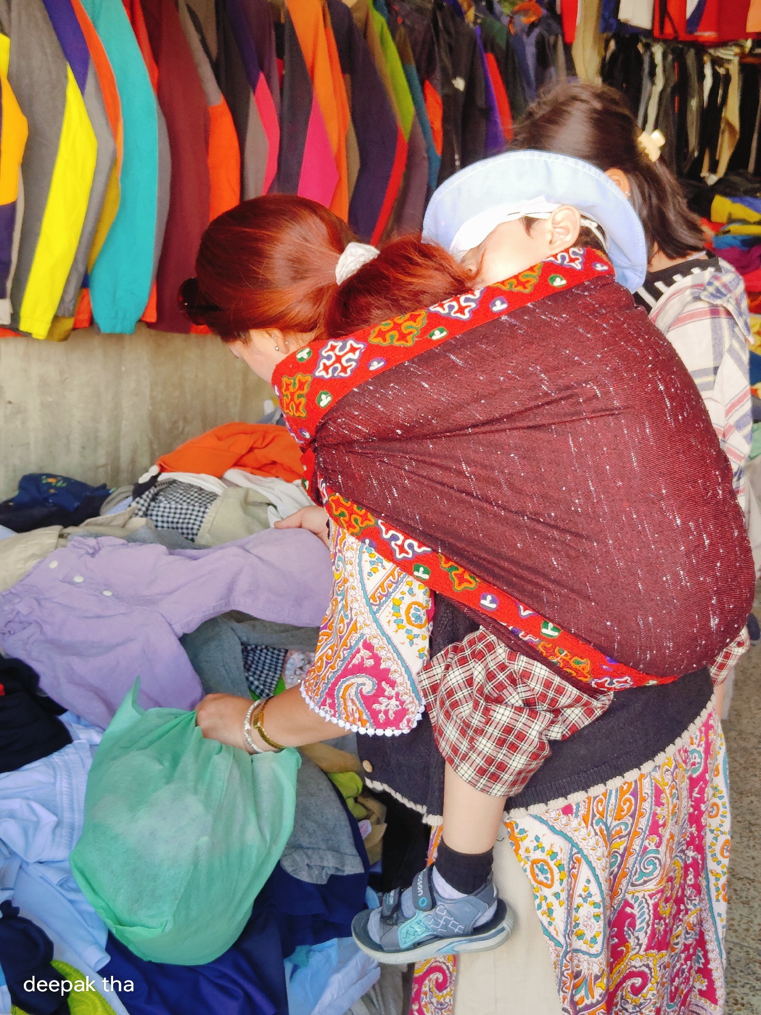 This is a medium shot photograph taken at a market in Leh, India. The central figure is a woman with reddish-brown hair, wearing a patterned dress and a dark, sequined wrap that cradles a baby. The baby, wearing a light blue hat, is partially visible, with its head resting on the woman's shoulder. The woman is holding a light green plastic bag in her right hand. In the foreground, there are piles of colorful clothing, including a purple shirt and a red and white plaid shirt. The background shows a stall with a variety of colorful garments hanging on display. The lighting is bright and diffused, suggesting an outdoor setting. The overall scene depicts a bustling market environment with a focus on the mother and child.