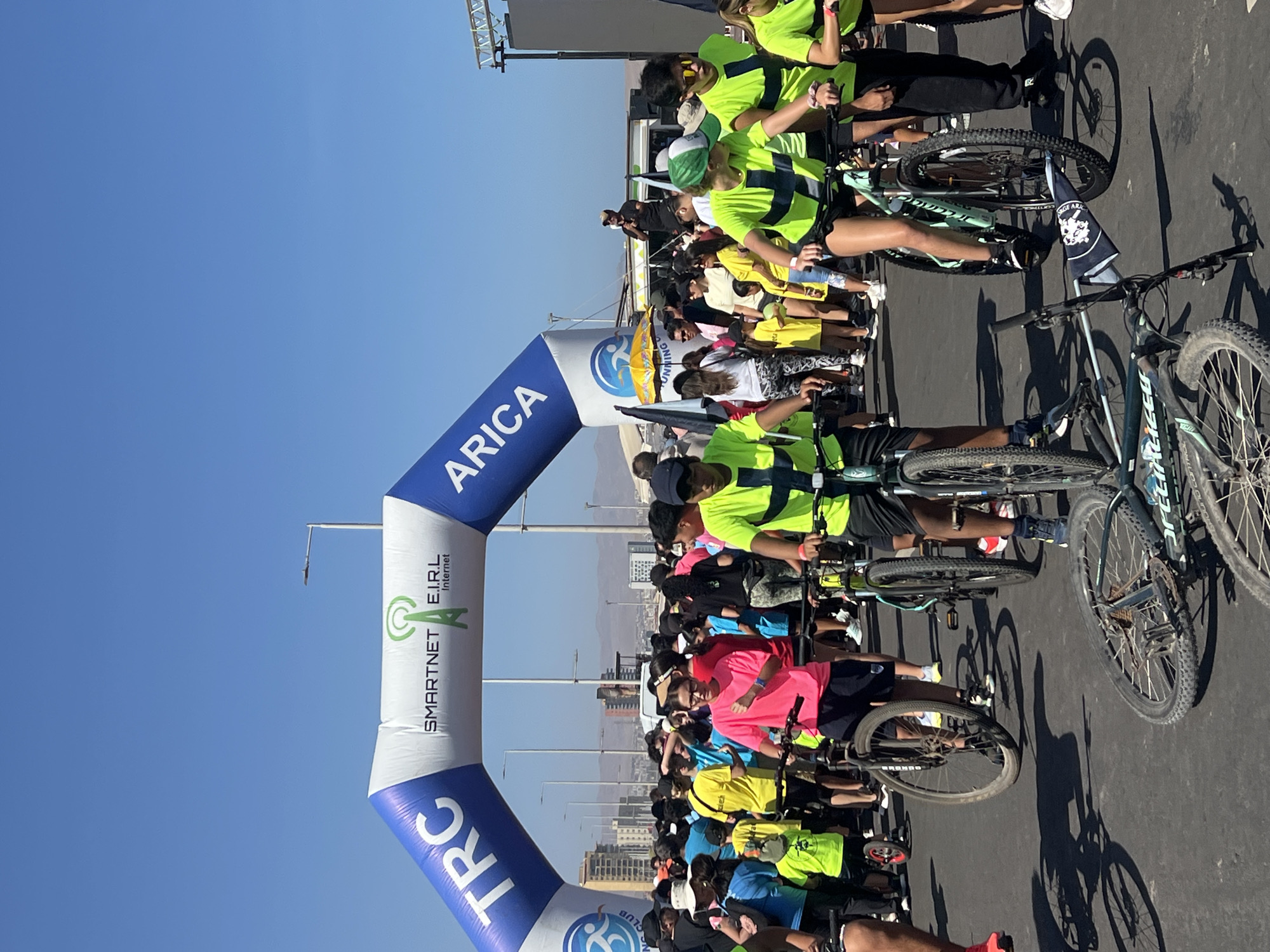 The image shows a cycling event in Arica, Chile. A large inflatable archway with "ARICA" and "SMARTNET E.I.R.L Internet" is visible at the starting point. Numerous cyclists, wearing various colored shirts, are lined up with their bicycles, ready to begin the race. Buildings and the city skyline are visible in the background, indicating an urban setting, and the clear blue sky suggests a sunny day. The event appears to be well-attended and organized, with a large crowd and clear markers. The bikes have different colors.