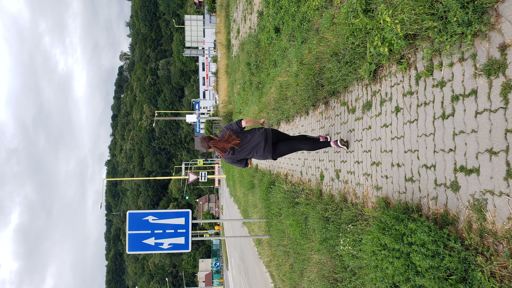 A woman with brown hair, wearing a dark gray hoodie and black leggings, runs away from the camera on a paved walkway. The walkway is bordered by lush green grass and weeds on both sides. In the background, a road with a traffic sign showing lanes for going straight and turning left is visible. Beyond the road, there are buildings and a heavily wooded hill under a cloudy sky. The scene appears to be in an urban or suburban area, possibly on the outskirts of Prešov, Slovakia, given the presence of European-style road signs and architecture.