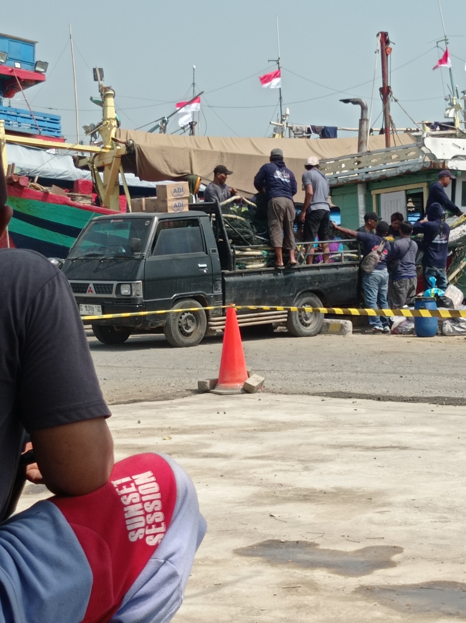 A group of people are gathered around a black pickup truck in what appears to be a harbor or dock area, with boats and flags visible in the background. The truck is loaded with what looks like fishing nets and other gear. Some individuals are standing, while others are sitting or leaning. One person in the foreground is wearing blue sweatpants with red accents and the text "SUNSET SESSION" printed on the thigh. The scene is lit by daylight, suggesting it is daytime. The environment suggests a working port, possibly in the city of Tegal, Indonesia, given the presence of Indonesian flags.