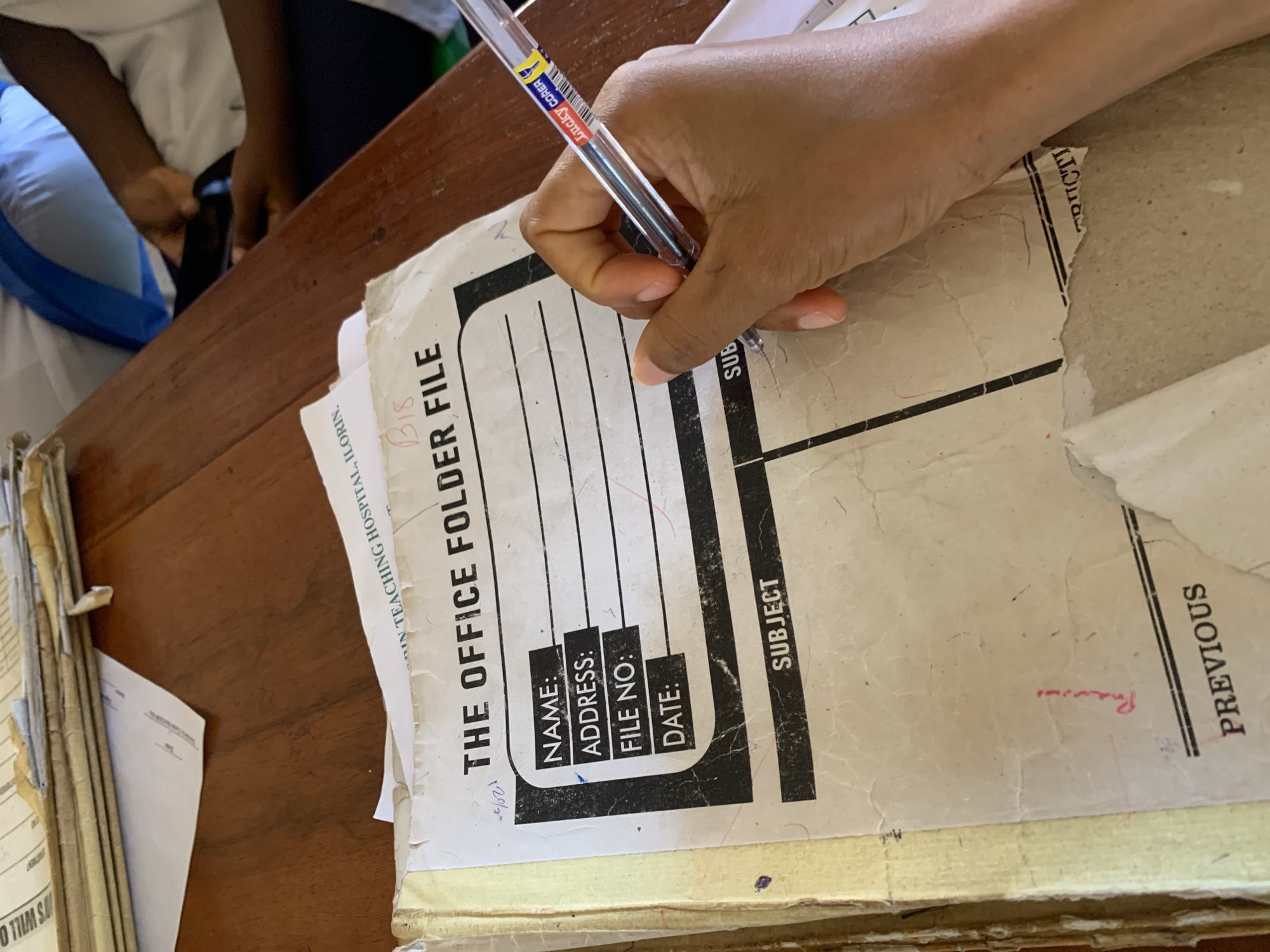 A person is filling out "The Office Folder File," which is a file folder for a patient from a Nigerian medical office or hospital. The file has the following fields to be filled out: "Name," "Address," "File No," "Date," and "Subject." The person is using a clear pen labeled "Lucky CORER." The office or hospital name is "University Teaching Hospital, Ilorin." The file is designated "B18". The word "previous" is written at the edge of the file. The scene is taking place in Apata Yakuba, Nigeria.