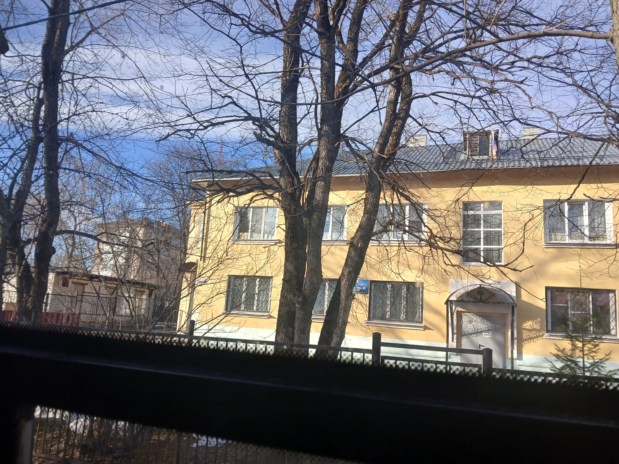A three-story yellow building is visible through bare trees. The building has a gray roof and white-framed windows. A door with a small awning is on the right side of the building. A fence runs along the front of the building. In the background, another building can be seen. The image is taken from inside a building, looking out through a window with a visible frame. A sign is visible on the building, partially obscured by the trees. The Russian flag is located on the roof. Based on the yellow building, likely a multi-apartment house, and the presence of Russian signage, it is most likely located in the city of Berezniki, Russia.