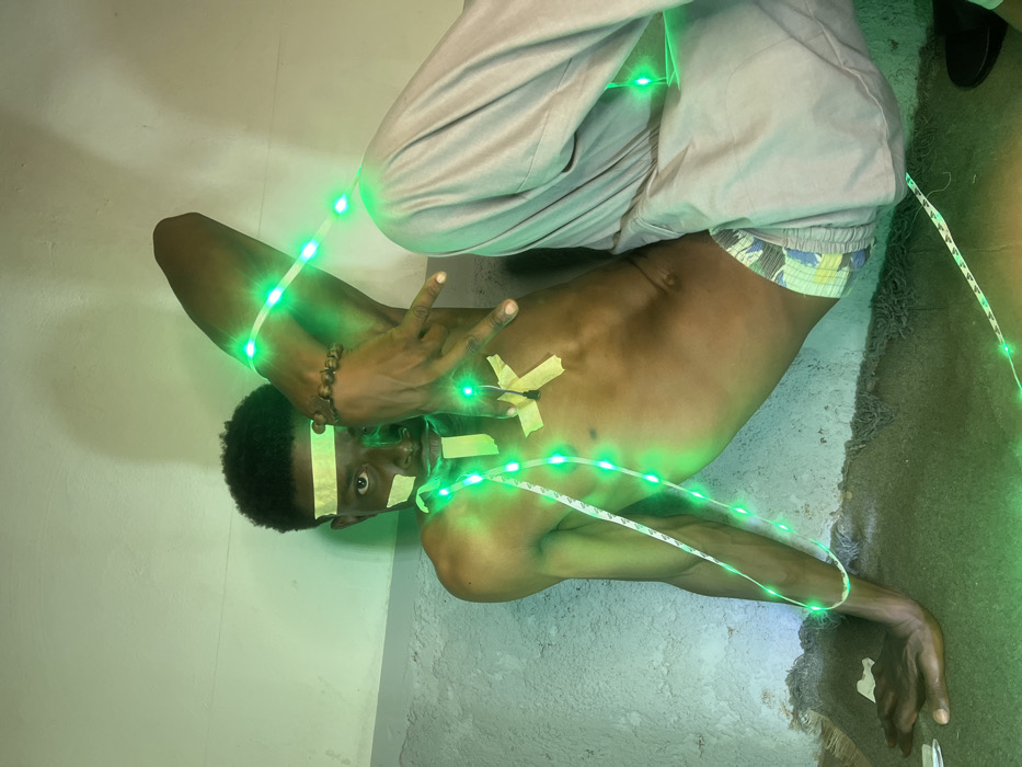 A male subject is depicted in an indoor setting within the city of Shen, Nigeria. He is positioned in a semi-crouched pose, leaning against a light-colored, textured wall. His torso is bare, displaying a lean physique. He wears light-colored trousers, with patterned boxer briefs visible at the waistline. A dark, beaded bracelet is on his right wrist. Multiple strips of yellow adhesive tape are affixed to his forehead, cheeks, and chest. A string of illuminated green LED lights is draped over his upper body, secured in places by the tape, and extends towards the dark, textured floor surface. His right arm is raised, with the hand open and fingers splayed. His left hand rests on the floor. The subject's gaze is directed forward, towards the camera.