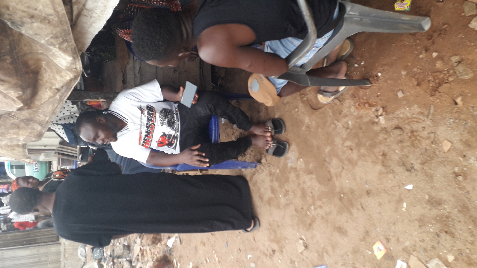 The image captures an outdoor scene in Onitsha, Nigeria. Several individuals are present: one man is seated on a blue plastic chair, holding a mobile phone. He is wearing a white t-shirt with black detailing and black pants, with black sandals on his feet. Another person is standing behind him, bending slightly. The ground is predominantly brown dirt, scattered with small rocks and pieces of debris. A grey plastic chair is visible in the background. There are some items on the right, on the ground, that are unidentifiable. To the left, there appears to be some makeshift coverings of cloth forming a rudimentary shelter. Overall, the scene is situated in a cluttered, potentially low-income area.