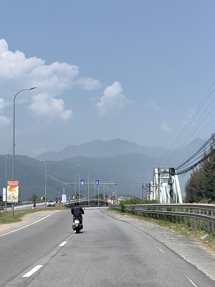 The image captures a highway scene in Hải Vân Ward, Vietnam, under a clear blue sky with scattered white clouds. A paved road, marked with white solid and broken lines, extends into the distance. A single individual, wearing a dark jacket, operates a white scooter in the center of the road, moving away from the viewer. To the right, a metal guardrail parallels the road, beyond which lies overgrown vegetation and pine trees. On the left, multiple streetlights line the roadside, along with a tall pole displaying a vertical, colorful billboard advertisement. Further down the road, a white car and other vehicles are visible approaching an overhead gantry structure with blue informational signs. To the right, a light blue metal truss bridge is prominent, with numerous black power lines and cables extending across the upper right portion of the frame and attached to structures near the bridge. A range of hazy, blue-grey mountains forms the background, spanning the entire horizon.