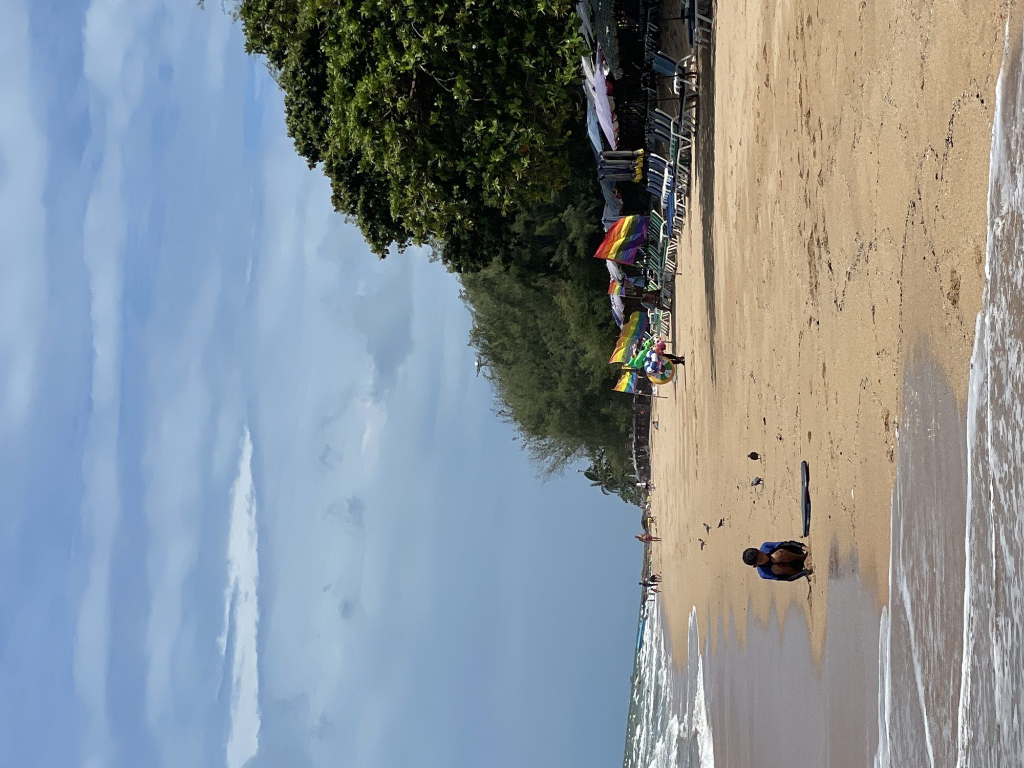 The image depicts a sandy beach with gentle waves washing ashore. A young person in a blue and black long-sleeved shirt is crouching on the wet sand near the waterline, appearing to play or examine something. A dark, flat object, possibly a boogie board or surfboard, lies on the sand next to them. Several birds are scattered on the beach in the mid-ground. Further down the beach, beach chairs and umbrellas are set up, and some people are walking or engaged in activities. Several rainbow-colored flags are visible, suggesting a festive or pride-themed event or location. The background features a line of trees and vegetation, with a cloudy sky overhead. The scene is consistent with a public beach in Pattaya City, Thailand, likely during daylight hours.