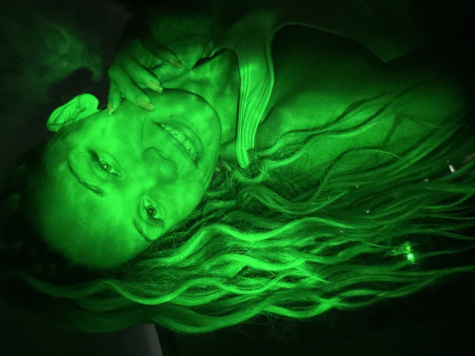 The image is a close-up, low-angle, indoor portrait of a woman illuminated by a bright green light. The woman is positioned on the left side of the frame, with her head tilted and her gaze directed towards the viewer. She has long, wavy, blonde hair that cascades down her left side. Her right hand is raised, touching her cheek and neck. She is smiling, revealing her teeth. The green light casts a strong, artificial glow over her skin, hair, and clothing, which appears to be a tank top. The background is dark and indistinct, suggesting an indoor setting with minimal ambient light. The overall mood is dramatic and somewhat otherworldly due to the intense green illumination. There is no discernible location in the city of Rusanovo, Russia visible in this image.