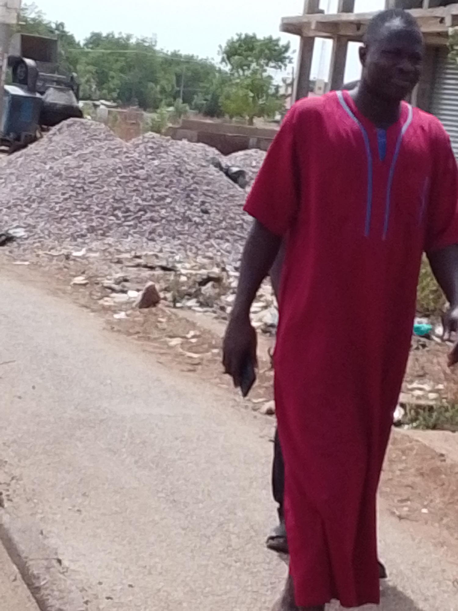 In Tamale, Ghana, a man is walking on a cracked asphalt road, adjacent to a large pile of gravel. He is wearing a long, maroon garment with blue trim around the neck. He is carrying a dark object, possibly a phone, in his left hand. Behind him, there is a piece of heavy machinery next to the gravel pile. In the background, trees are visible, along with a building under construction.