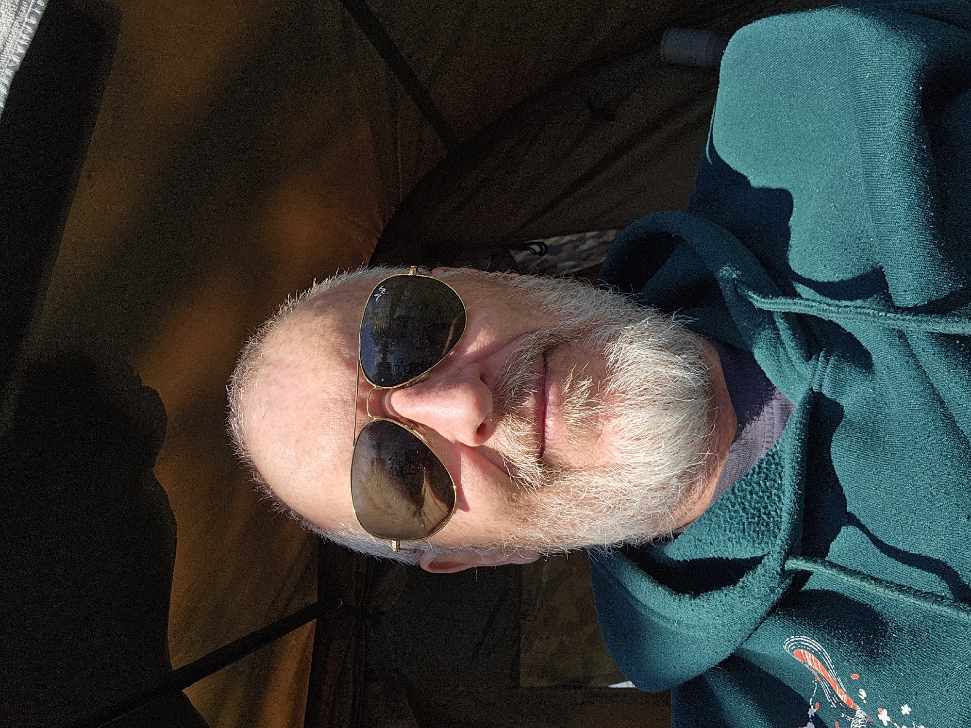 A close-up, rotated image shows an older man with a bald head and white beard wearing gold-rimmed sunglasses. The man is wearing a dark green hooded sweatshirt. The background appears to be the interior of a tent or shelter, with the tent walls in a brown color. The lighting suggests an outdoor environment with direct sunlight. The location, Gavisse, France, cannot be confirmed as there are no identifiable landmarks or location markers in the image.