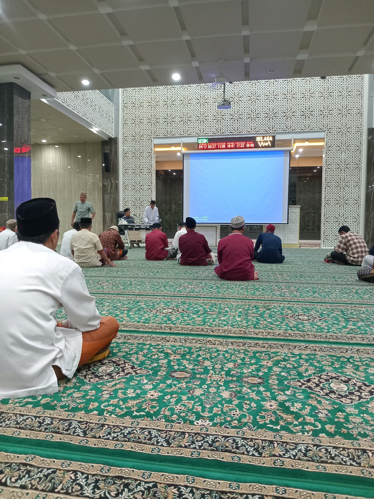 In Randuagung, Indonesia, within a mosque, a group of men are gathered on a green carpet with intricate patterns. They are seated on the floor facing a large projector screen displaying a blue image. A few individuals are standing near the screen, where a projector is mounted on the ceiling. The mosque features patterned walls and columns. Some of the men are wearing traditional Islamic attire, including caps. The atmosphere appears calm and attentive, suggesting a religious gathering or presentation.