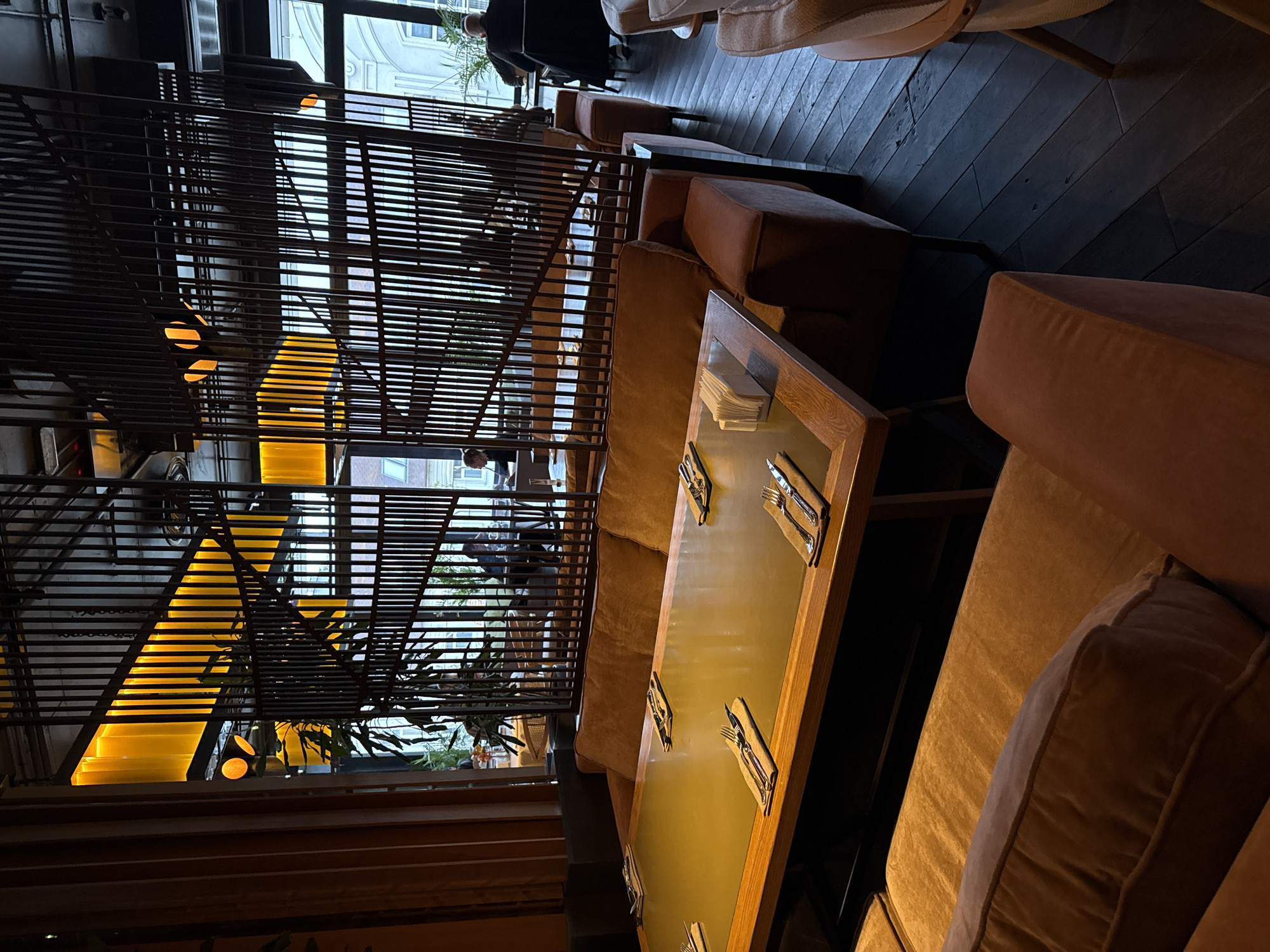 This image captures the interior of a dimly lit, modern restaurant or cafe, presented in a warm, inviting atmosphere. The scene features rows of plush, upholstered sofas or banquettes in a rich brown or tan color, positioned alongside rectangular glass-topped tables with wooden frames. Each table is neatly set with cutlery (forks and knives) placed on napkins, suggesting readiness for service.
A prominent decorative element is a tall partition constructed from numerous thin, horizontal dark slats, which visually separates the foreground seating area from the background. Beyond this partition, a striking architectural feature with bright yellow or amber backlighting, possibly a staircase or ramp, adds a vibrant glow to the space. Through the gaps in the partition and large windows on the far side, several indistinct figures, presumably patrons, can be seen seated or moving about.
The flooring consists of dark wooden planks. The lighting is predominantly artificial and warm, casting a soft glow that implies it is evening or nighttime. Through the windows, hints of urban buildings with classic architectural details are visible, consistent with a European city like Kazan, Russia. No readable text is visible anywhere in the image. The overall impression is one of a stylish, comfortable, and possibly upscale dining establishment.