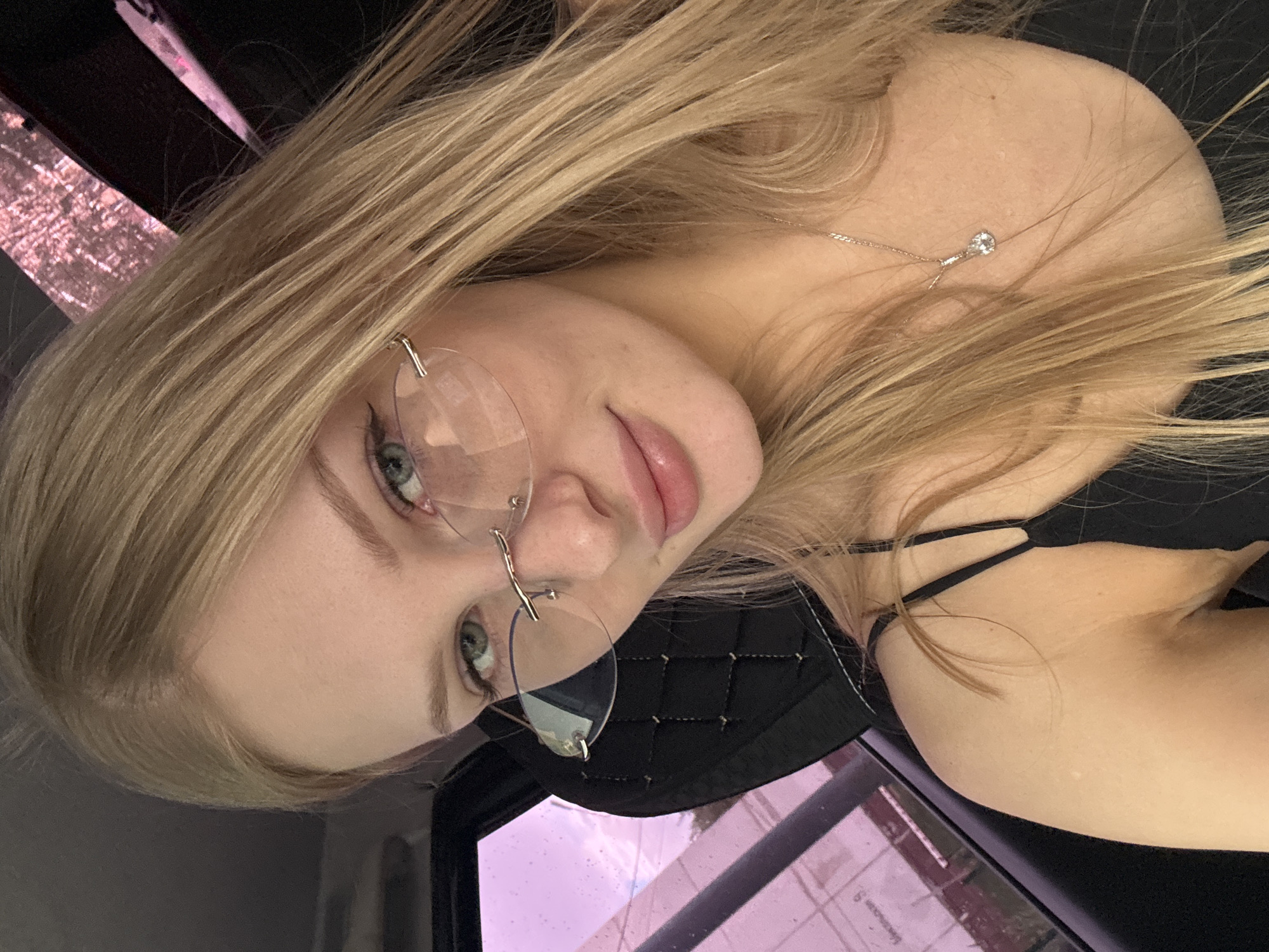 A young woman with blonde hair and light blue eyes is shown in a close-up, sideways shot. She is wearing rimless glasses with clear lenses and a thin silver necklace with a small diamond pendant. Her lips are slightly parted, and she appears to be looking at the camera. Her hair is long and falls around her face and shoulders. She is wearing a black top with thin straps visible. In the background, there is a black quilted material and a pinkish-purple tinted surface that appears to be a window or screen. The overall lighting suggests it might be daytime, but the setting is unclear. The image was taken in Perm, Russia.
