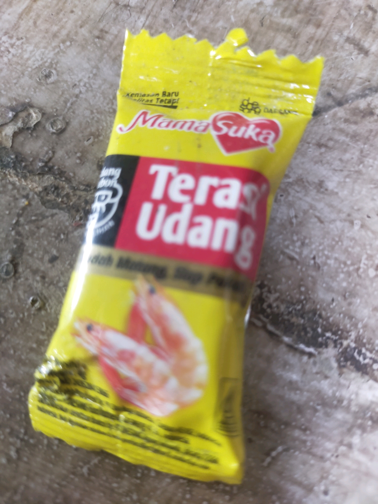A close-up, blurred shot shows a yellow packet of "Mama Suka" shrimp-flavored seasoning on a textured, greyish-brown surface, possibly a wooden table with a worn finish. The packet features a prominent red rectangle with the words "Teras Udang" in white, indicating it is shrimp crackers or seasoning. A stylized red heart with the word "Suka" inside is also visible. The packet has jagged edges at the top. Two blurred images of pink shrimp are depicted on the lower part of the packet. The background is out of focus, but appears to be a neutral, textured surface. No people or discernible landmarks related to Mojodanu, Indonesia, are present.