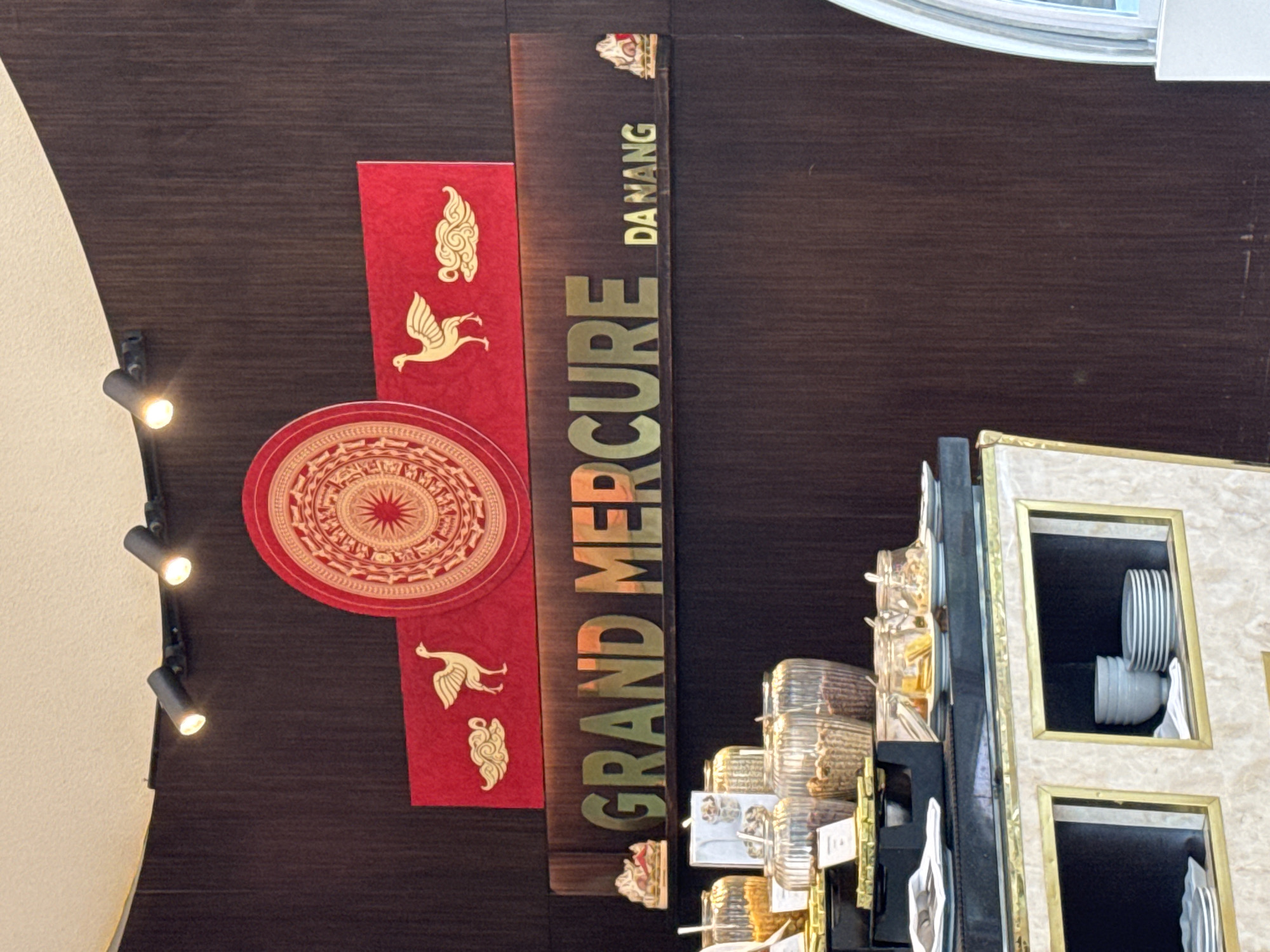 The image depicts a sign for the "Grand Mercure Da Nang" hotel in Da Nang, Vietnam. The sign is mounted on a dark brown wall or ceiling. The words "GRAND MERCURE" are vertically aligned in gold lettering, next to a vertical red banner that features two golden bird silhouettes and a red circular emblem. "DA NANG" is vertically aligned next to "MERCURE," with a small golden emblem above. To the right of the sign are decorations housed in framed cabinets with plates. Track lighting is visible to the left of the sign.